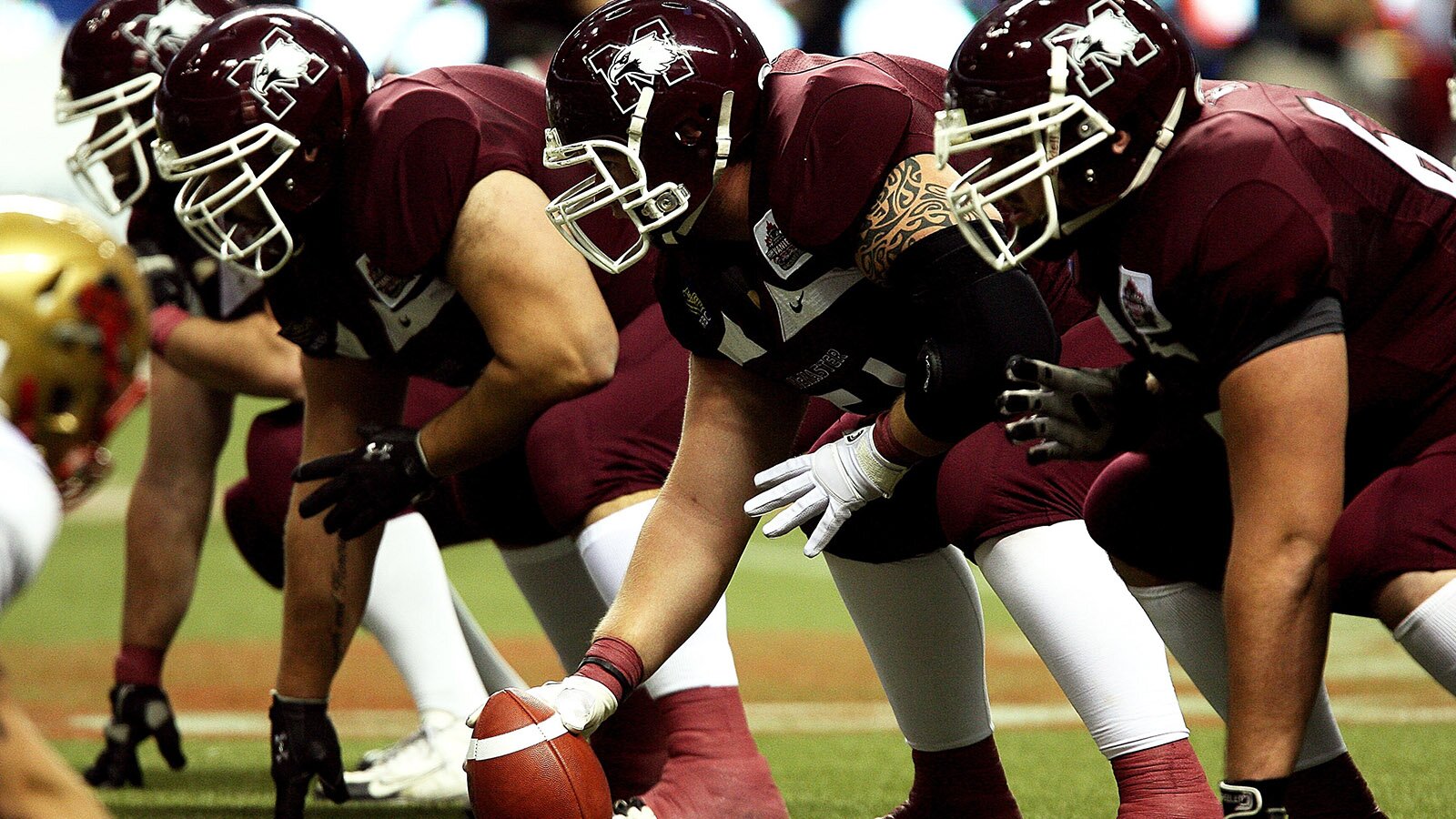 Zdjęcie przedstawia czterech zawodników rugby, którzy stoją na ugiętych kolanach i pochylają się nad murawą. Wszyscy ubrani są w ciemnobrązowe koszulki z krótkim rękawem, ciemnobrązowe, obcisłe spodenki do kolan, białe skarpety, sportowe buty, rękawiczki i ochraniacze. Na głowach mają czarne kaski ochronne z metalowymi, ażurowymi osłonami na twarzy.