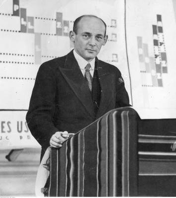 Fotografia przedstawia łysiejącego mężczyznę w średnim wieku ubranego w garnitur koszulę i krawat podczas przemówienia do ludzi.