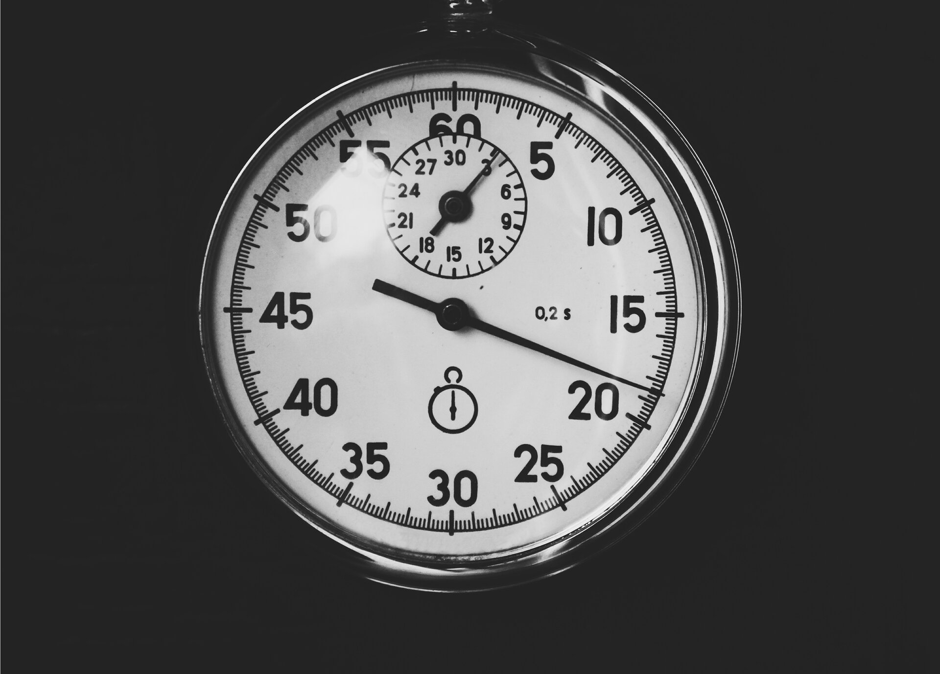 Na ilustracji okrągły stoper w srebrnej delikatnej obudowie sfotografowany z bliska, na czarnym tle. Ma białą tarczę, na której są cyfry i kreseczki zapisane na dwóch okręgach, jeden w drugim. Na zewnętrznym znajdują się liczby, na samej górze  - 60, następne liczby zwiększają się o 5 aż do 55, po którym znów następuje 60. Pomiędzy każdą z liczb zaznaczone jest po 5 średnich kresek, między którymi z kolei jest po 5 najkrótszych kresek. Liczbę 60 od dołu zasłania drugi mniejszy licznik z małą wskazówką i liczbami zapisanymi na okręgu. Na samej górze umieszczona jest liczba 30, kolejne liczby wzrastają o 3, aż 27 dotrze znów do 30. Końcówka dużej głównej wskazówki zatrzymała się na wartości 18,5, z kolei mała wskazówka  wewnętrznego licznika - na 3.