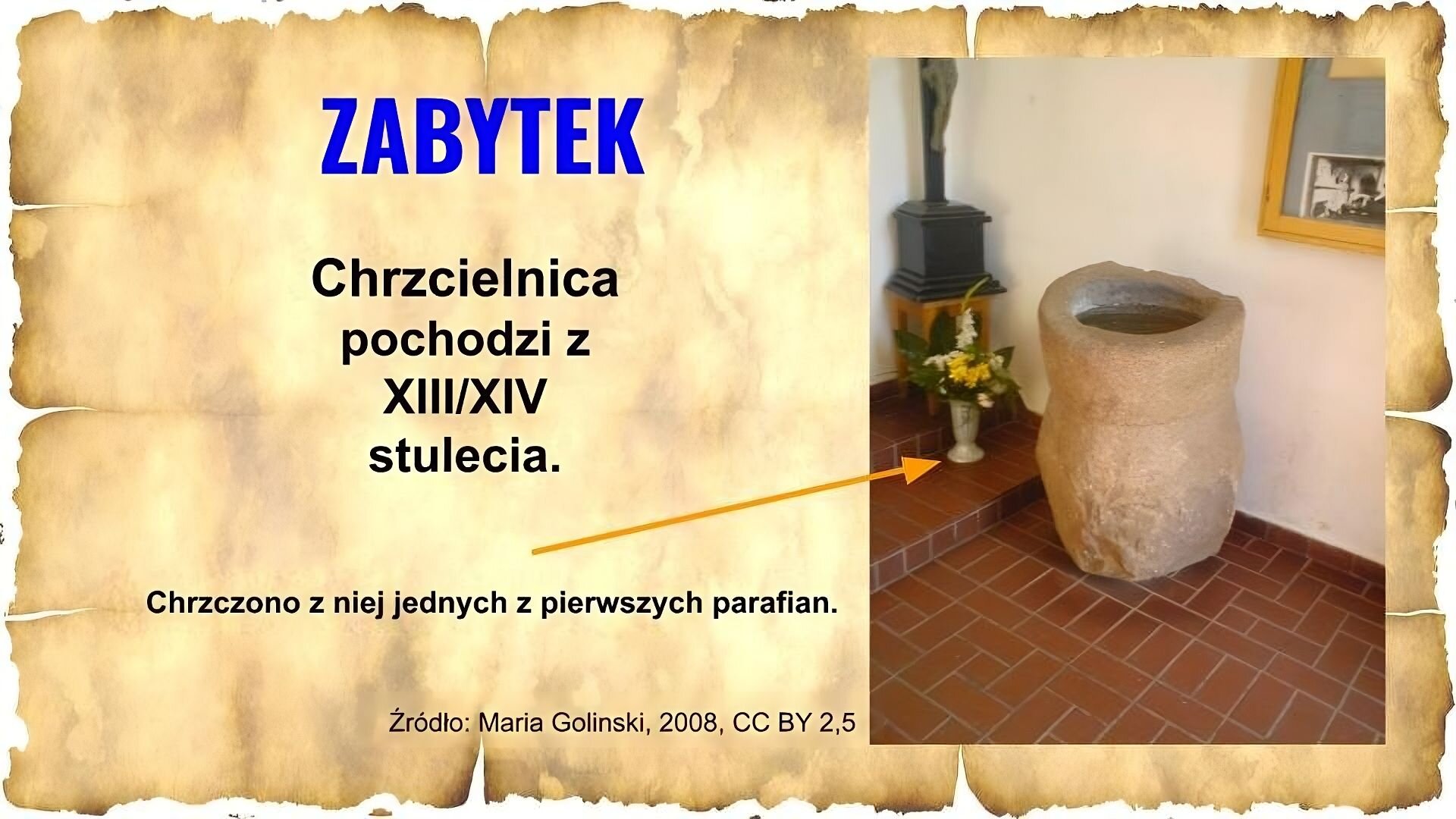 Slajd zawiera napis tytułowy: Zabytek. Poniżej jest napis: chrzcielnica pochodzi z XIII/XIV stulecia. Po prawej znajduje się zdjęcie, na którym przedstawiono chrzcielnicę – około metrowy kamienny dzban, który stoi na posadzce w pomieszczeniu. Obok niego stoją kwiaty, na ścianie za chrzcielnicą widać obraz. Przy ilustracji umieszczono napis ze strzałką wskazującą na strzelnicę: Chrzczono z niej jednych z pierwszych parafian. Na dole znajduje się napis: Źródło: Maria Golinski, 2008, CC BY 2,5.
