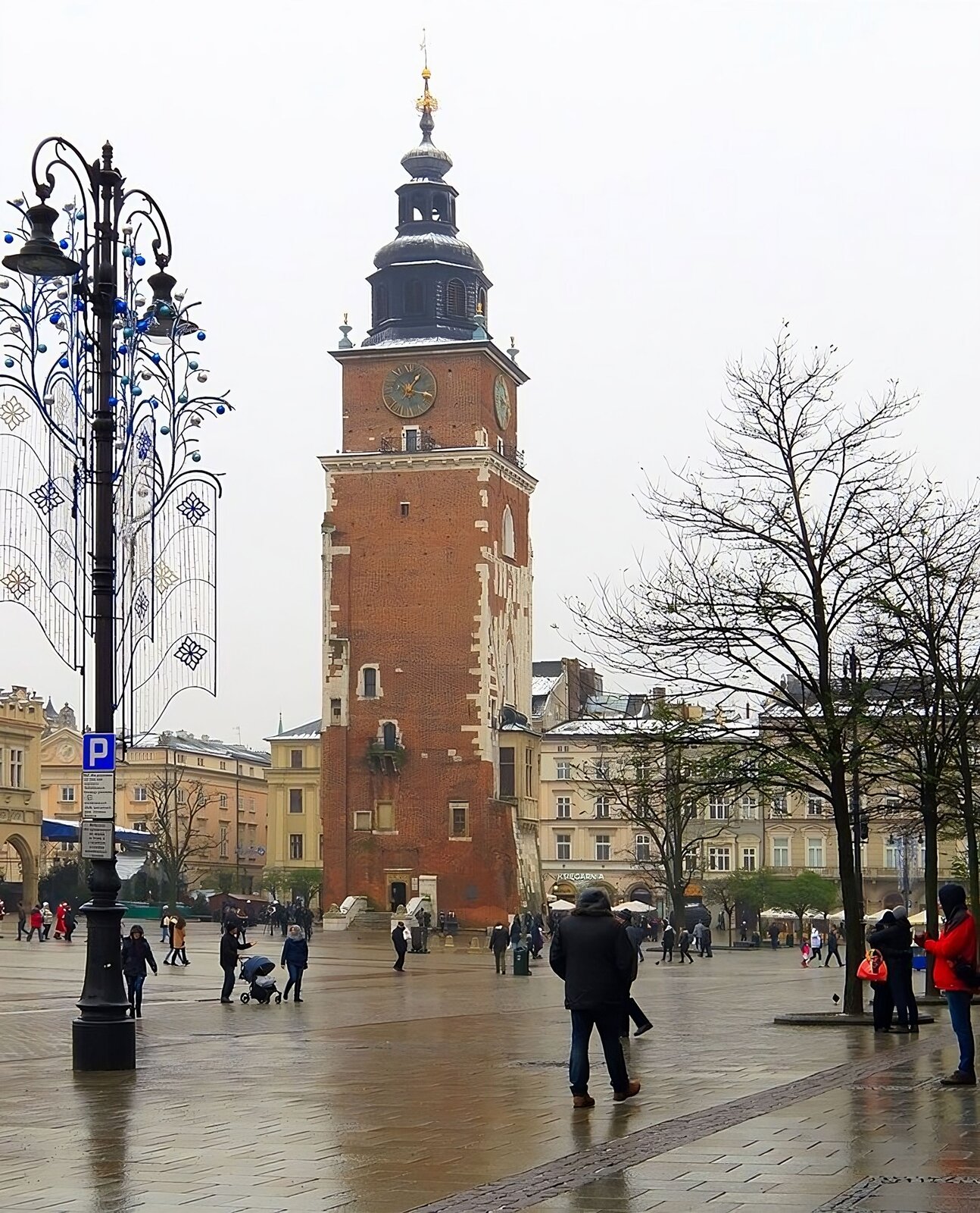 Zdjęcie przedstawia widok na wieżę nieistniejącego ratusza na rynku w Krakowie. Wieża o podstawie kwadratu, z wąskimi prostokątnymi oknami wyrasta ponad linię zabudowy miejskiej dookoła. Po jej prawej stronie widoczny na kondygnacji niewielki wykusz. Całość konstrukcji wzniesiona z cegły, z kamiennymi uzupełnieniami w ścianach i obramowaniach okien. Zwężenie w szczytowej części kondygnacji, prowadzące do wieży dzwonnej. Pod kopułą wieloboczną wieży na prezentowanych ścianach ratusza widoczna są tarcze zegarów. Zdjęcie wykonane w okolicach świąt Bożego Narodzenia, o czym świadczą miejskie dekoracje na pierwszym planie oraz bezlistne drzewa. 