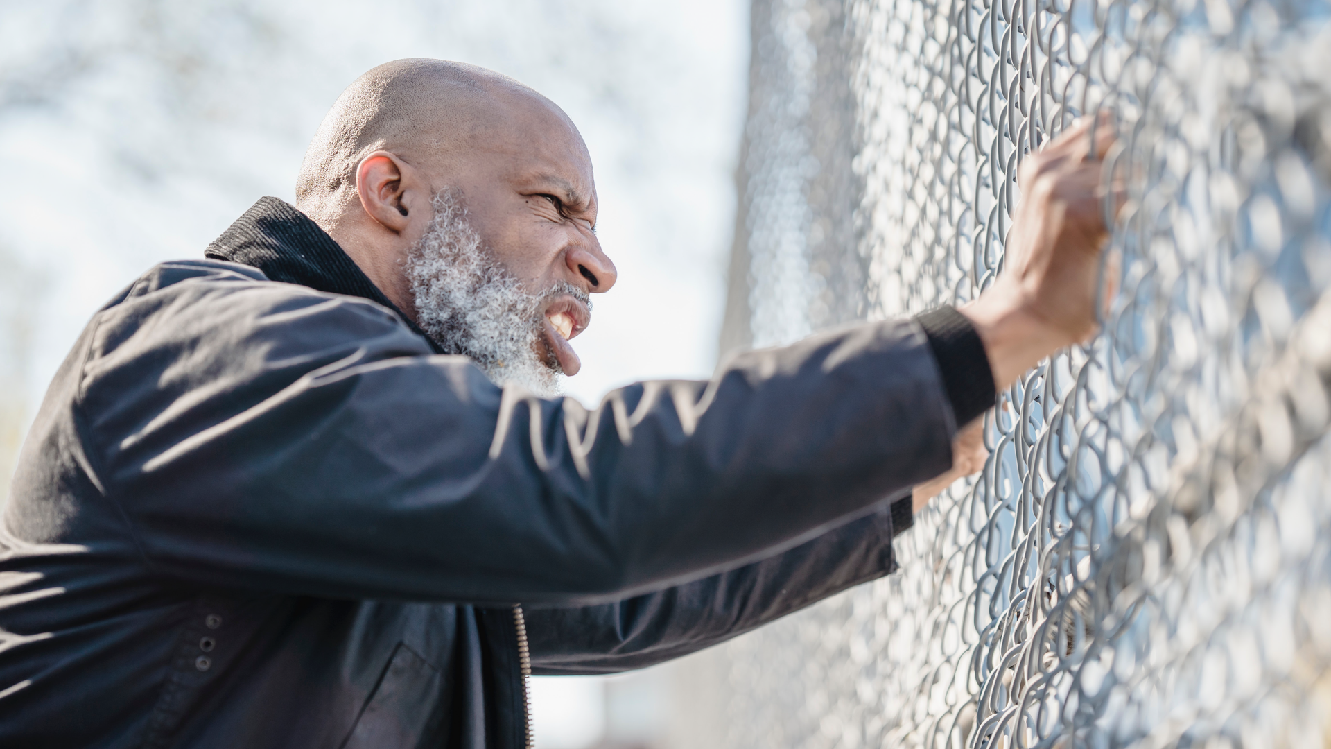Zdjęcie przedstawia łysego, starszego mężczyznę z brodą, ubranego w skórzaną kurtkę. Mężczyzna obiema rękoma chwyta za metalową siatkę ogrodzenia. Na jego twarzy widać grymas złości.