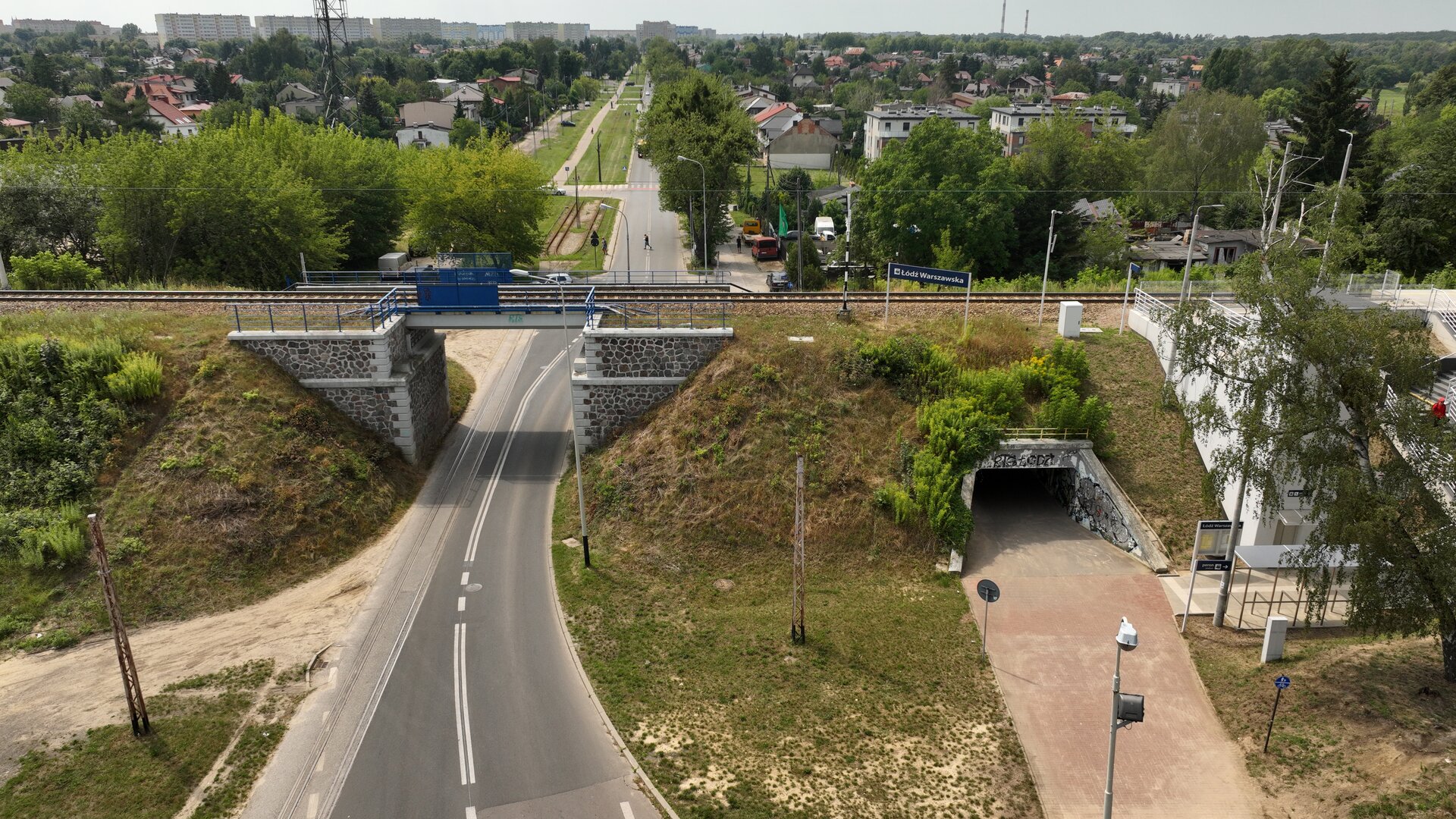 Ilustracja przedstawia przejście pod torami. Widoczny jest wiadukt nad drogą. Przez nasyp kolejowy obok wiaduktu poprowadzone jest przejście pod torami.