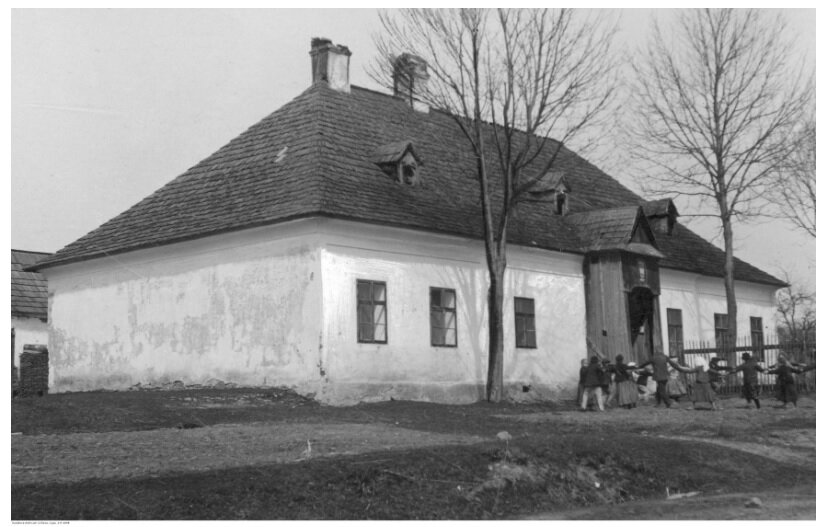 Fotografia przedstawia duży budynek ze spadzistym dachem, na którym widać dwa kominy. Przed budynkiem biegają dzieci i stoją drzewa.