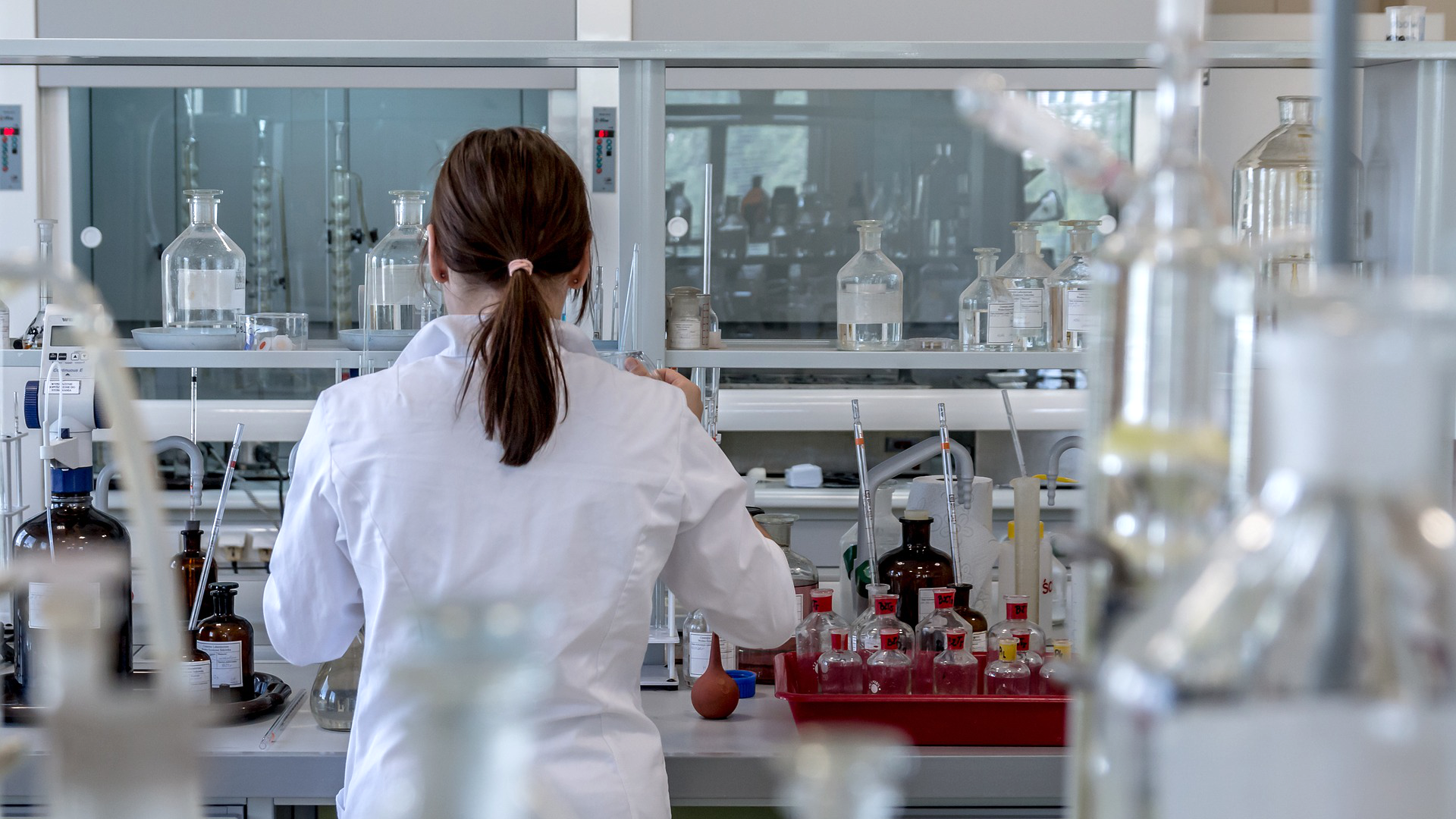 Zdjęcie przedstawia laboratorium. Na zdjęciu widoczne są szklane naczynia do wykonywania badań. Tyłem do kadru stoi kobieta w białym fartuchu.