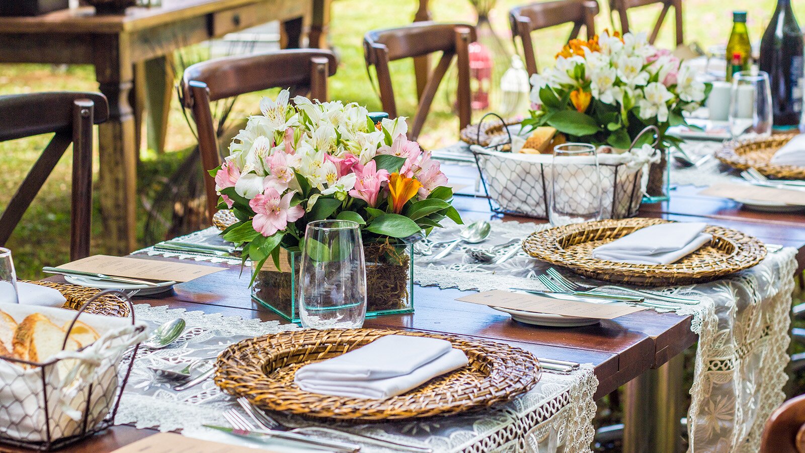 Zdjęcie przedstawia zastawiony stół na świeżym powietrzu, a na około niego ustawione krzesła. Główną dekoracją stołu są świeże kwiaty.