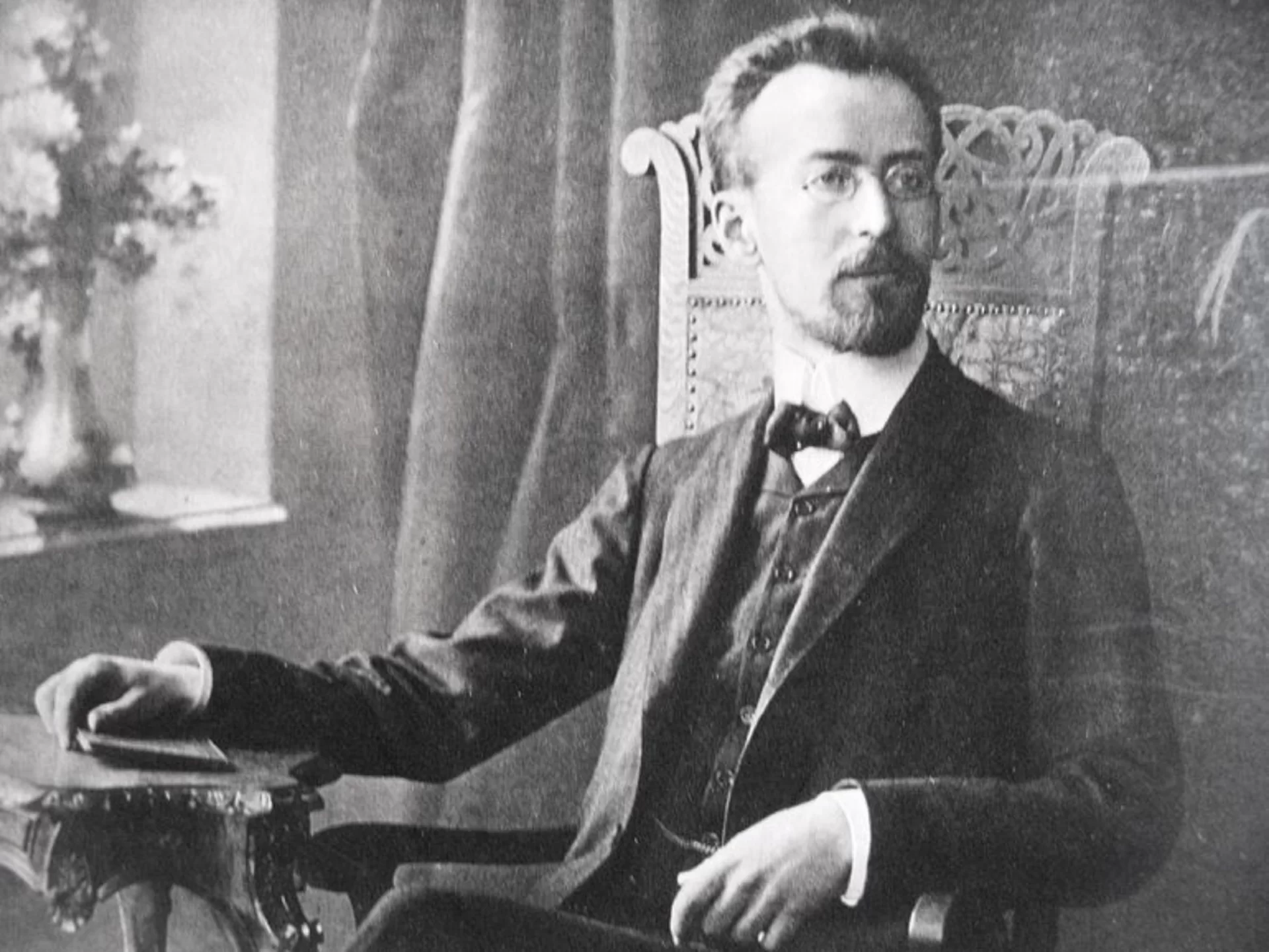 Ilustracja przedstawia portret Mieczysława Karłowicza - polskiego kompozytora i dyrygenta. Na foto jest widoczny szczupły mężczyzna siedzący na drewnianym krześle przy stoliku, w dłoni trzyma notes. Mężczyzna ma ciemne, gęste i zaczesane do tyłu włosy. Wyraz twarzy spokojny. Ubrany w białą koszulę, muszkę i garnitur.