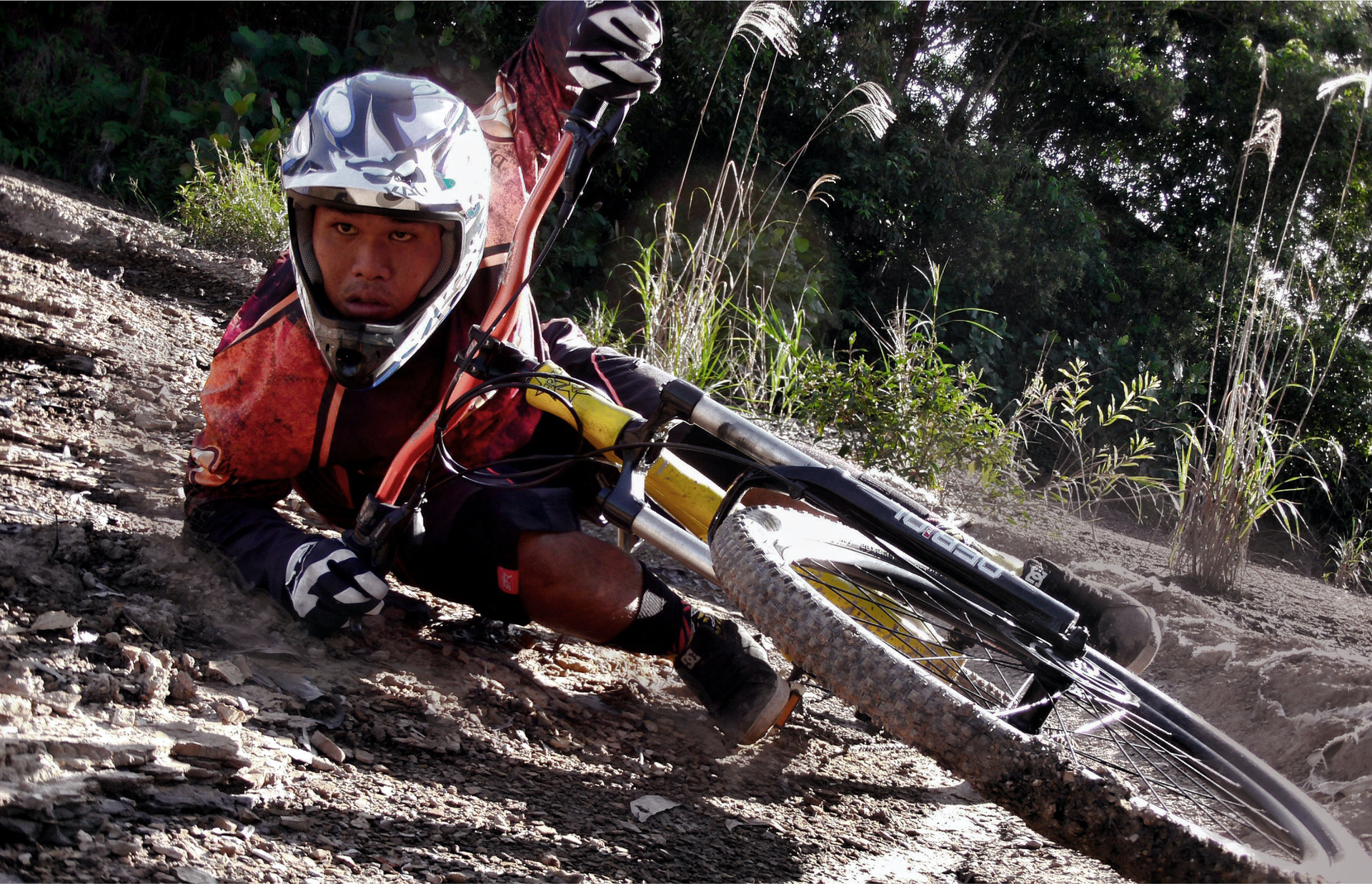 Zdjęcie przedstawia mężczyznę ubranego w kask i kombinezon do jazdy rowerem w terenie. Mężczyzna przewrócił się i leży na ziemi. Na nim leży rower.
