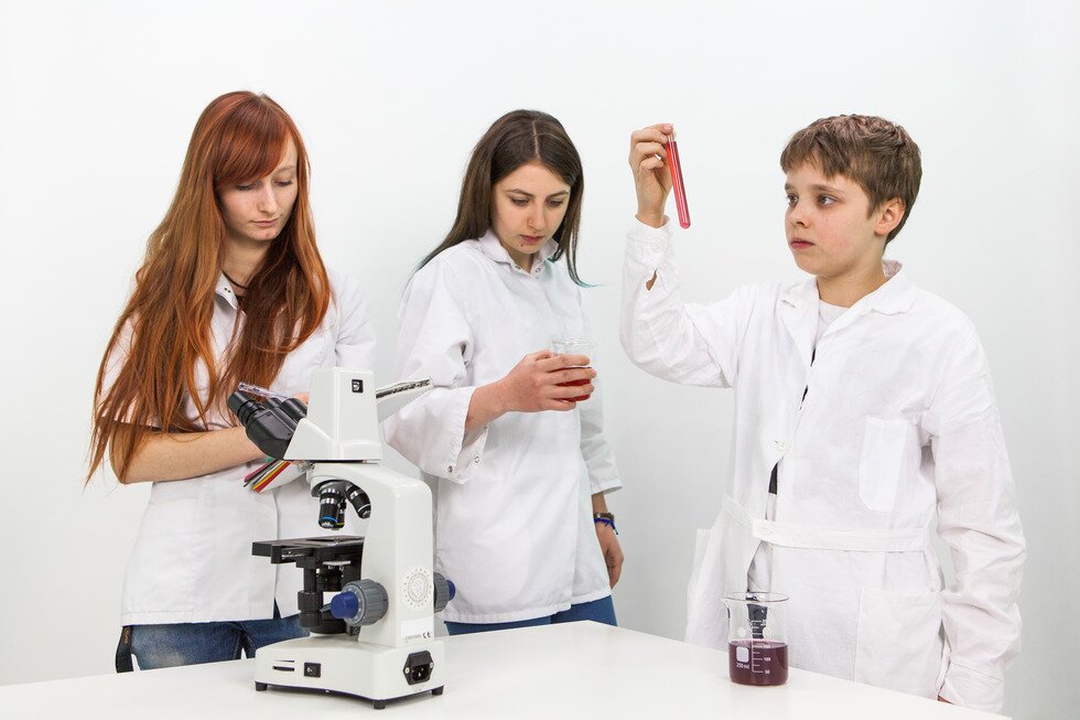 Zdjęcie młodzieży, dwóch nastolatek i jednego nastolatka stojących przy stole laboratoryjnym. Ubrani są w laboratoryjne, białe fartuchy. Na stole stoi mikroskop oraz zlewka z płynną substancją. Jedna z nastolatek trzyma w ręku zlewkę z płynną substancją i się jej przygląda. Druga nastolatka robi notatki w zeszycie. Nastolatek trzyma w ręku probówkę wypełnioną substancją ze zlewki i przygląda się jej.