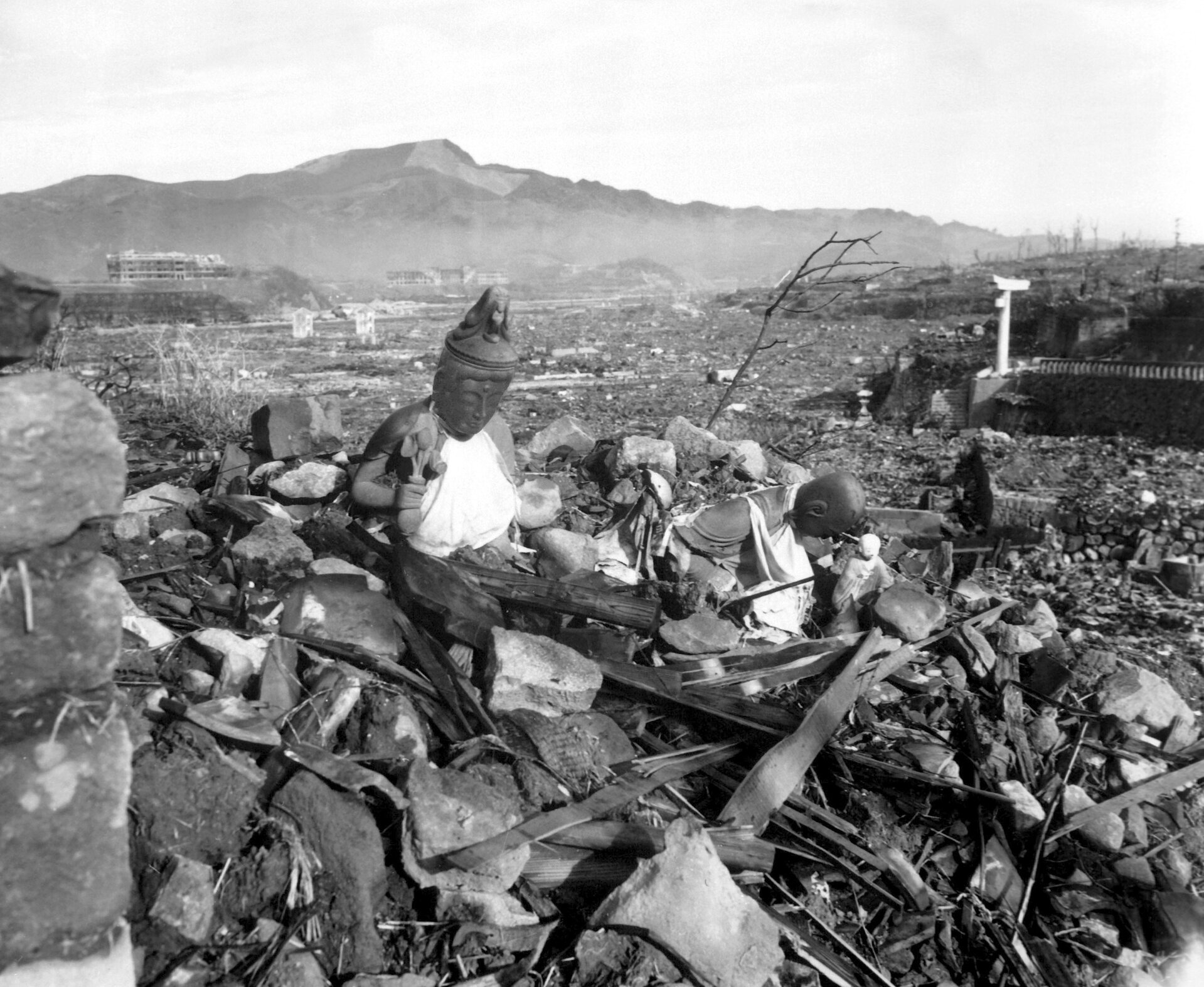 Zdjęcie przedstawia zniszczone przez wybuch bomby atomowej tereny Nagasaki. Fotografia czarno‑biała. Na pierwszym planie widoczne sterta gruzu, połamanych desek i konstrukcji oraz fragment figurki Buddy. Nieco dalej widać ogrom zniszczeń spowodowanych przez wybuch wojny. Prawie wszystko zostało zrównane z ziemią. W oddali widać szkielety budynków. W tle zarys gór.