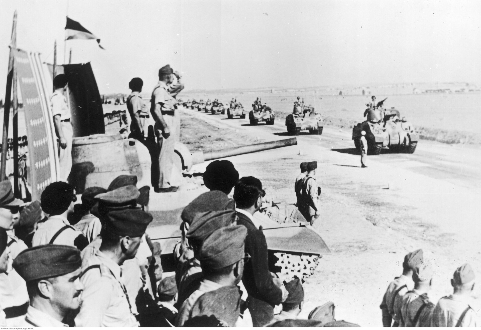 Zdjęcie przedstawia defiladę Samodzielnej 2 Brygady Pancernej przed generałem Kazimierzem Sosnkowskim. Drogą jeszcze szereg czołgów w równych odstępach. Generał Sosnkowski salutuje przejeżdżającym żołnierzom, stojąc na czołgu. W jego pobliżu są licznie zgromadzeni polscy wojskowi.