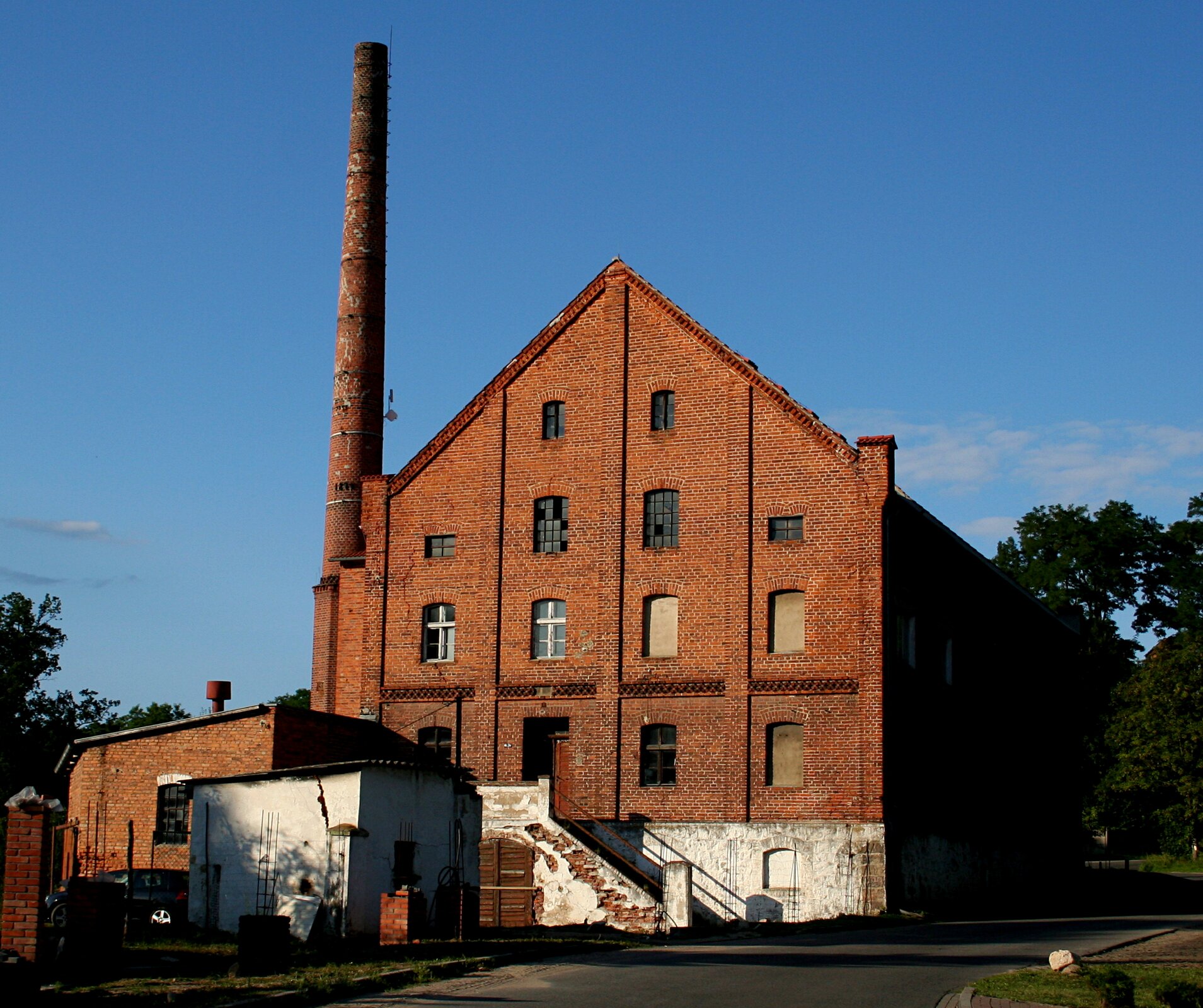 Zdjęcie przedstawia kilkupiętrowy ceglany budynek, przy którym stoi wysoki komin. Do budynku prowadzą schody. W tle widoczne jest bezchmurne niebo.
