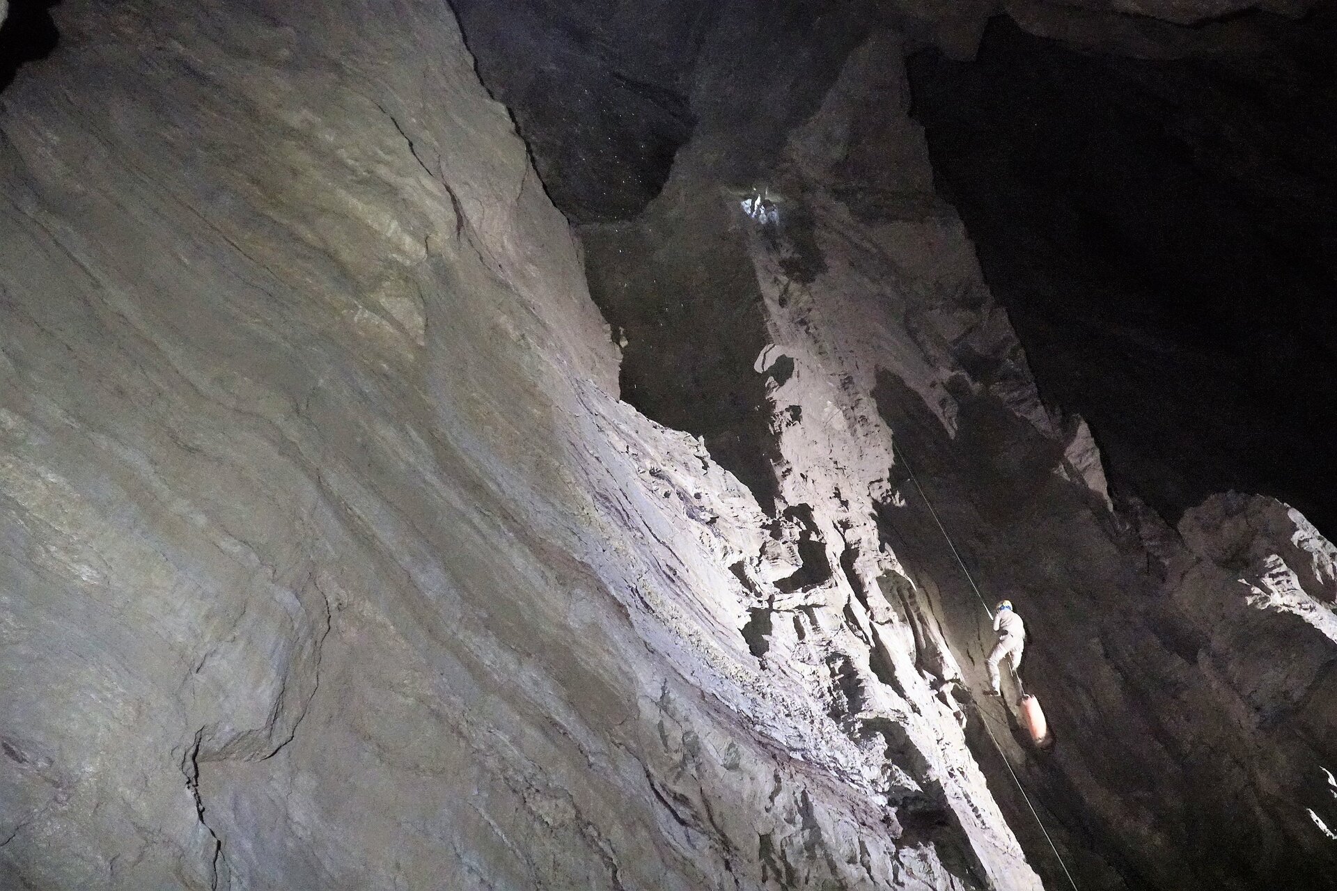 Na zdjęciu znajduje się wnętrze jaskini. Po lewej stronie jest potężna ściana skalna. PO środku znajduje się lina, po której zjeżdża wgłąb jaskini postać. 