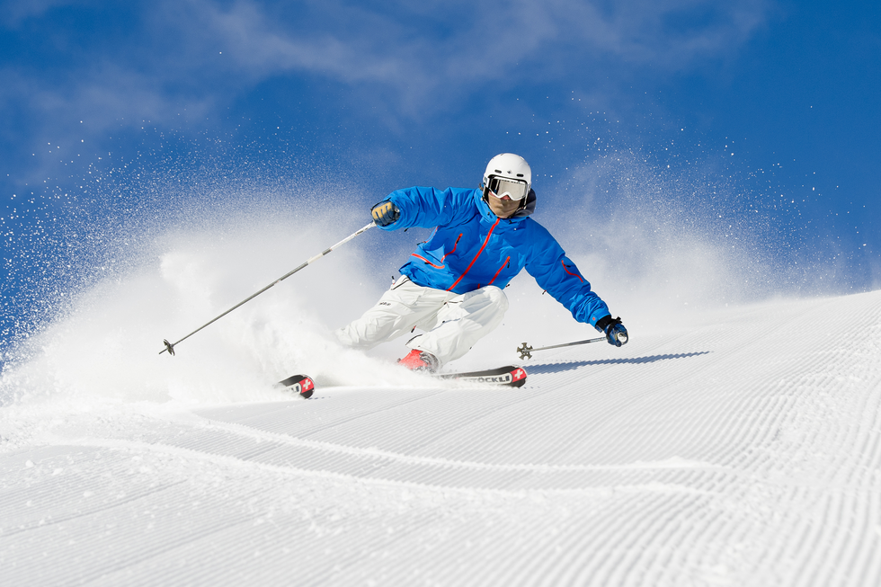 Fotografia narciarza na stoku. Widać śnieg i bezchmurne niebo. Narciarz ubrany jest w kask i gogle przeciwsłoneczne oraz kombinezon narciarski. W dłoniach trzyma kijki narciarskie.