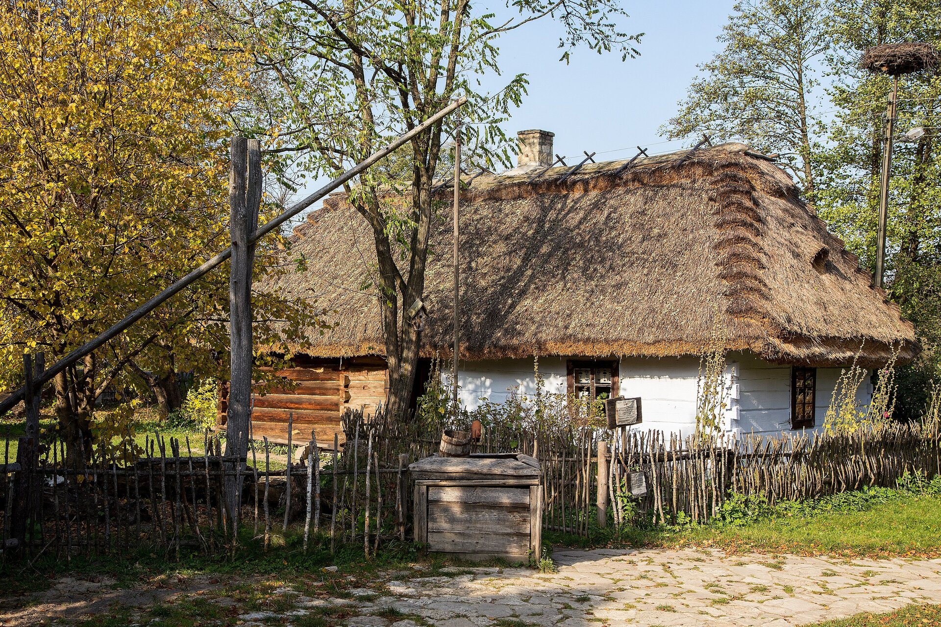 Na zdjęciu stara chatka z bielonymi ścianami. Pokryta strzechą. Obok studnia i drewniany płot. Dookoła wysokie drzewa. 