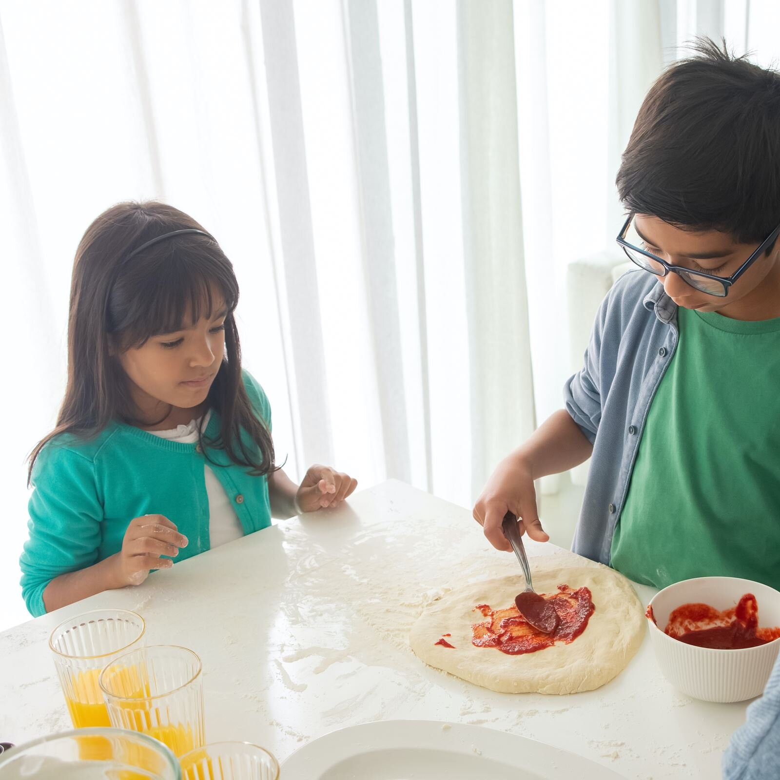 Zdjęcie przedstawia dziewczynkę i chłopca w wieku szkolnym, którzy stoją przy białym blacie i przygotowują pizzę. Chłopiec, stojący po prawej stronie, w prawej ręce trzyma łyżkę, którą rozsmarowuje sos pomidorowy na placku pizzy. Na lewo od pizzy stoi miska z sosem pomidorowym. Dziewczynka, stojąca po lewej stronie, przygląda się jego pracy. Na stole widoczne są też szklanki z sokiem pomarańczowym, fragment talerza.