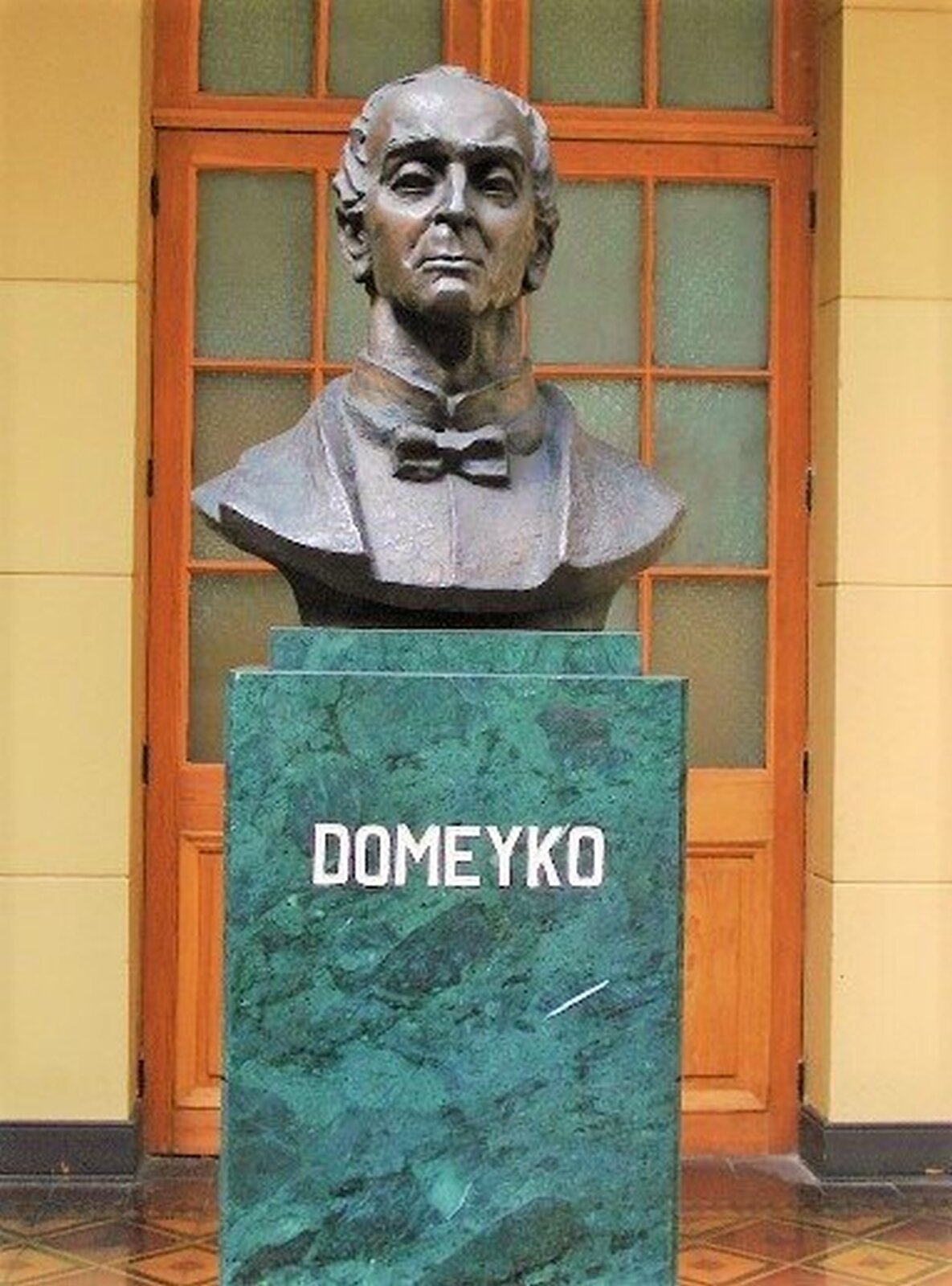 Fotografia przedstawia wykonane z brązu popiersie Ignacego Domeyki. Popiersie ustawione na marmurowej bryle na której widnieje złoty podpis Domeyko. W tle drewniane drzwi.