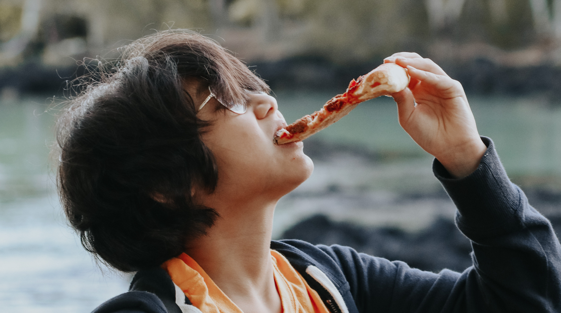 Zdjęcie przedstawia chłopca z włosami do ramion i w okularach. Chłopiec ma lekko uniesioną głowę i je kawałek pizzy.