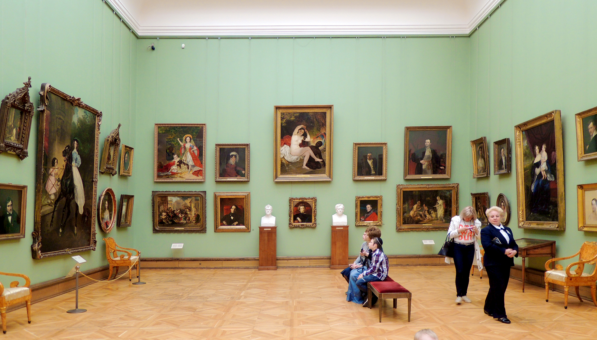 Ilustracja przedstawia wnętrze muzeum. Na ścianach wiszą różnej wielkości obrazy w ramach. Przy największej ścianie stoją na podwyższeniach dwa popiersia. Pośrodku znajduje się ławeczka, na której siedzą podziwiający eksponaty ludzie.