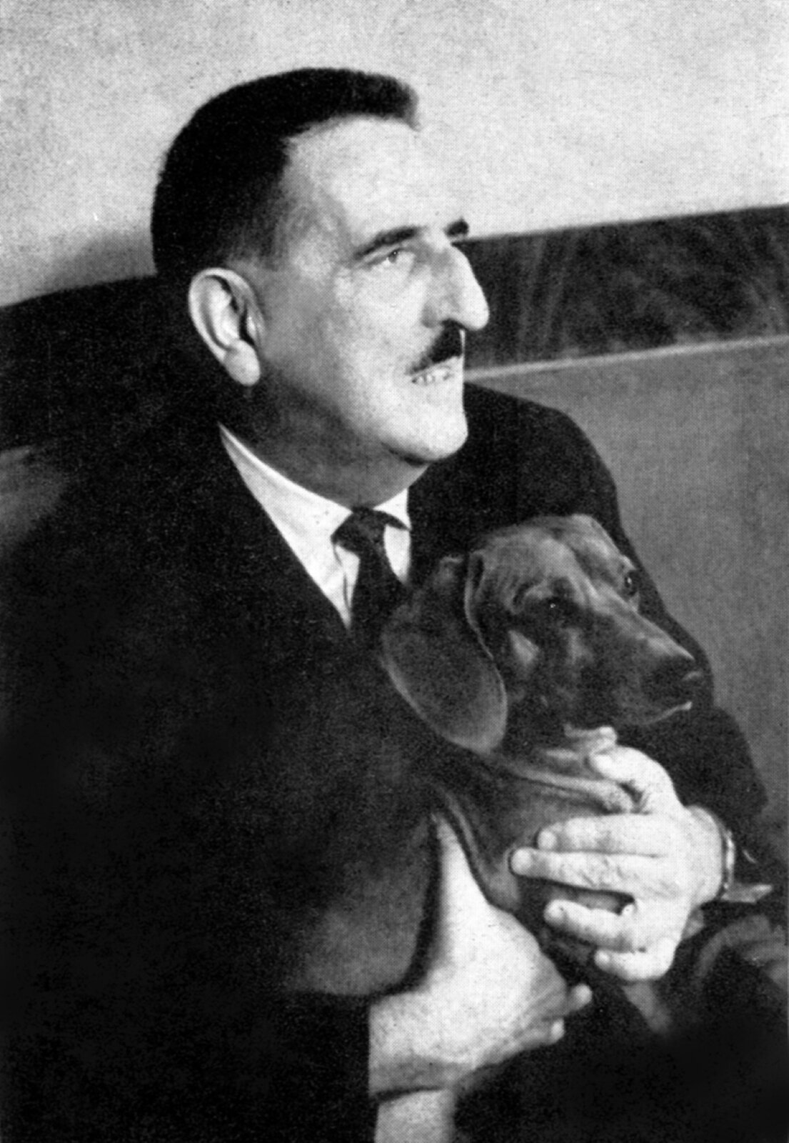 Ilustracja przedstawia czarno‑białe zdjęcie. Na zdjęciu znajduje się siedzący mężczyzna spoglądający na lewą stronę. Na kolanach obiema rękami trzyma jamnika. Mężczyzna ma czarne włosy i wąsy. Długi nos i wyciągnięta w dół brodę oraz muskularne uszy. Jego usta są delikatnie rozchylone. Ma na sobie czarną marynarkę pod którą, znajduje się biała koszula z czarnym krawatem. 