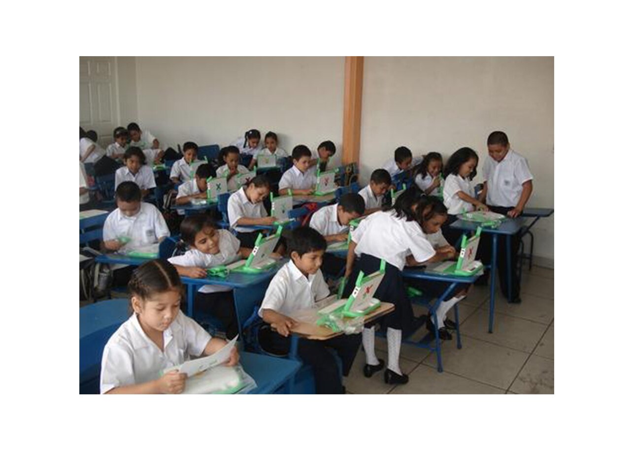 Klasa szkolna. W pojedynczych ławkach siedzą około 10-letnie dzieci, dziewczynki i chłopcy. Dzieci mają przed sobą szkolne laptopy, w które patrzą. Jedna z dziewczynek czyta to, co jest napisane na kartce.