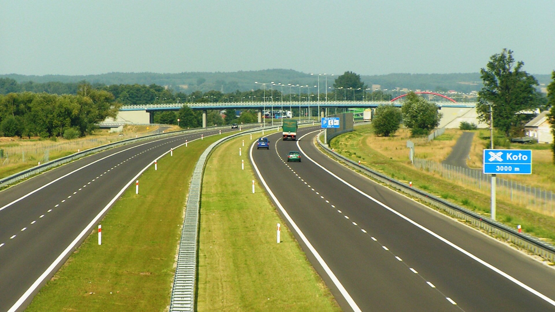 Fotografia przedstawiająca fragment autostrady A2, pojazdy poruszają się po dwóch jezdniach oddzielonych pasem zieleni, w tle wiadukt nad autostradą.