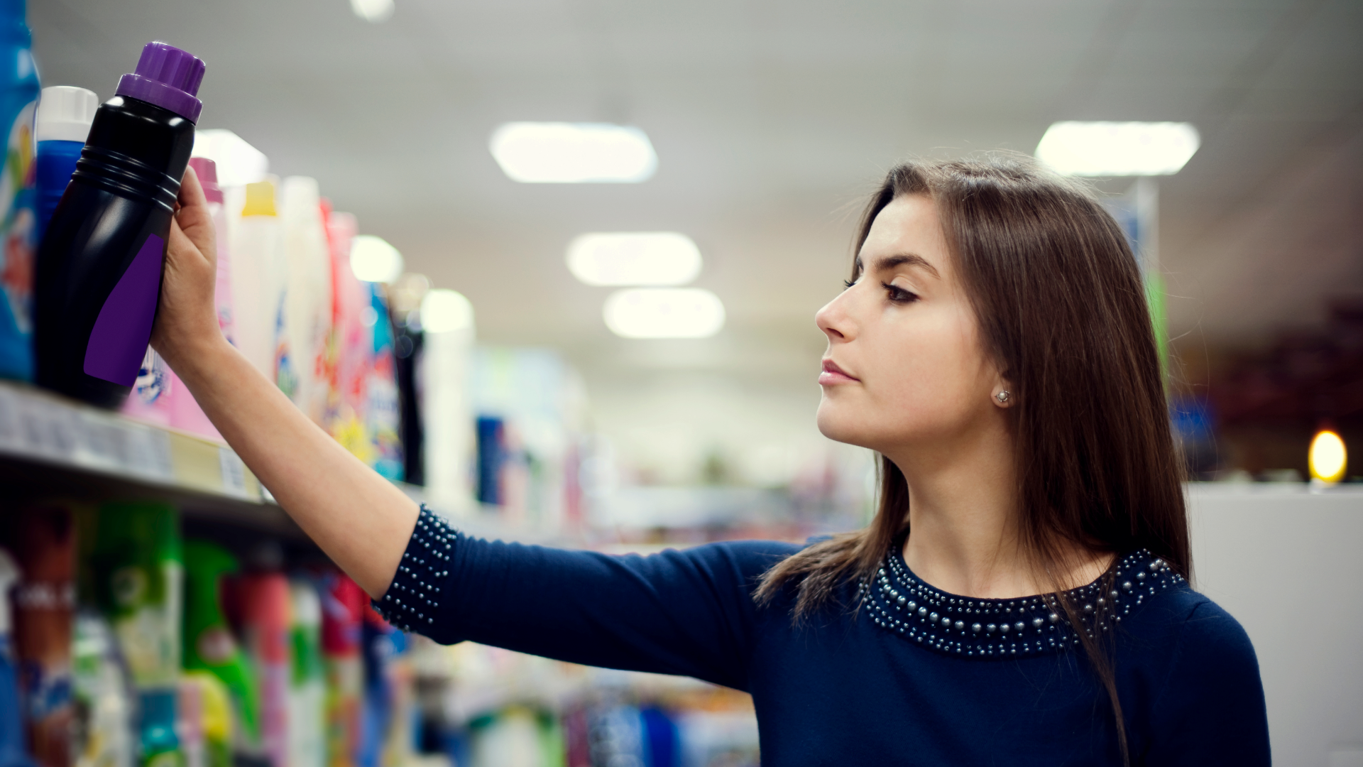 Zdjęcie przedstawia młodą kobietę sięgającą prawą ręką po butelkę ze środkiem czystości stojącym na półce w sklepie.