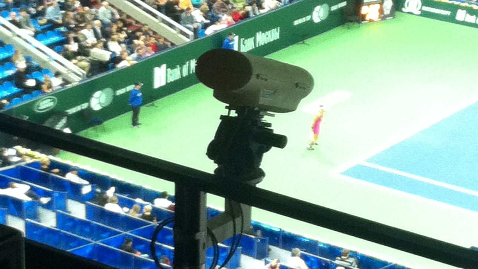 Zdjęcie przedstawia kamerę, która zamocowana jest na barierce i skierowana na kort tenisowy widoczny poniżej. Wokół kortu widoczne są kaskadowo ustawione rzędy niebieskich siedzeń. Na części z nich siedzą ludzie.