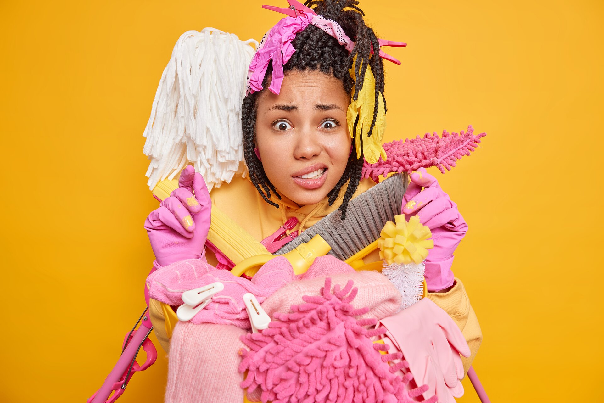 Zdjęcie przedstawia kobietę ze zdezorientowaną miną, która na dłoniach ma lateksowe rękawiczki. O jej ramiona są oparte dwa mopy, a przed nią leży sterta ściereczek, szczotek oraz spinacze do prania.