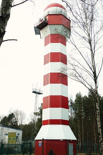 Na zdjęciu wieża radaru meteorologicznego. Pomalowana jest w biało‑czerwone poziome pasy. Ma średnicę kilku metrów, zbudowana jest na planie wielokąta. U podstawy i na górze nieco szersza. W górnej części wieży umiejscowione również dwa małe balkony. Wieża u szczytu przybiera kształt kuli, dookoła jej nasady znajduje się taras zabezpieczony barierkami. Konstrukcja ma wysokość około kilkunastu metrów, jest nieco wyższa od pobliskich drzew.