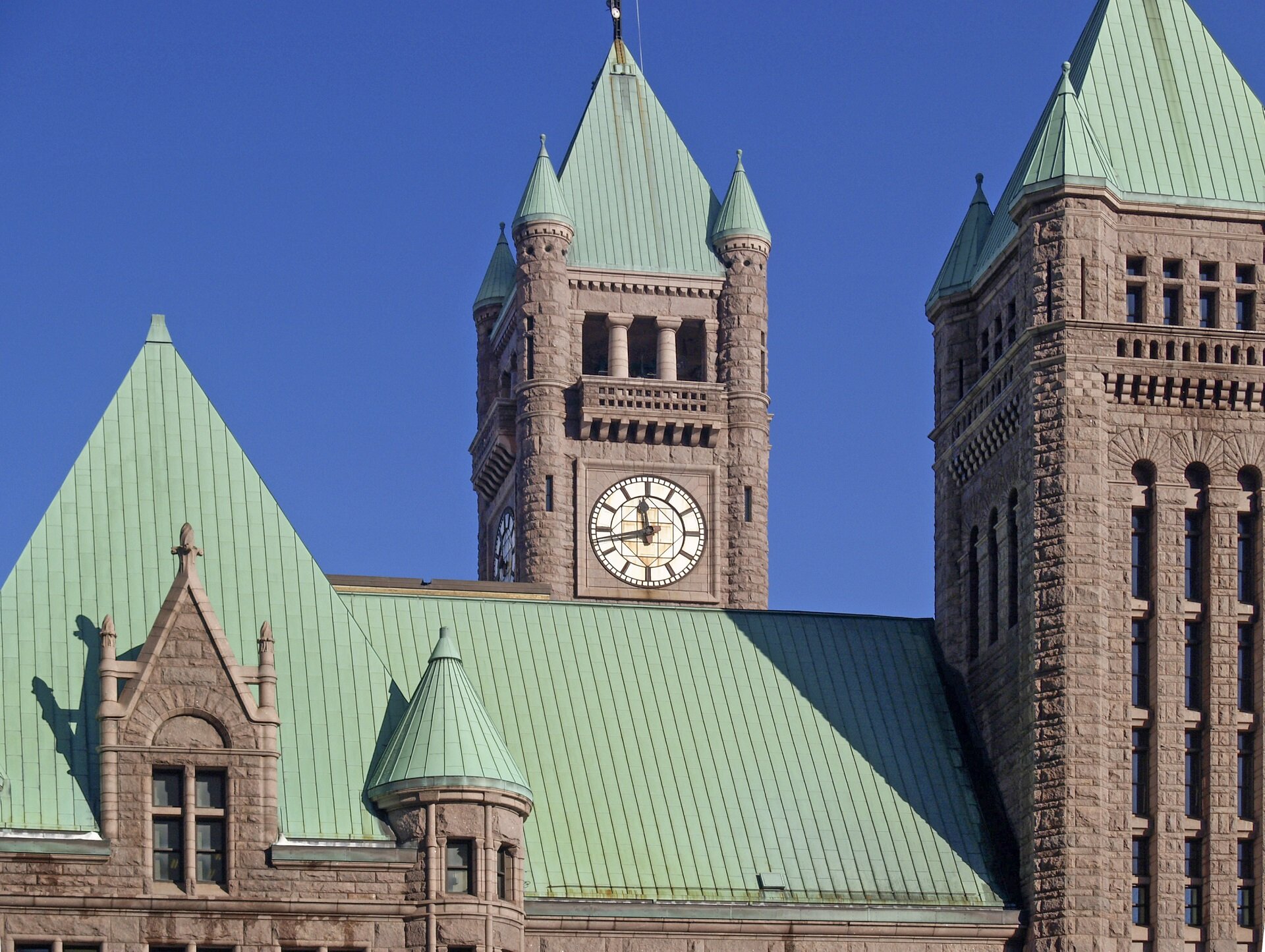 Zdjęcie przedstawia dach Urzędu Miasta w Minneapolis. W centralnej części grafiki oraz z jej prawej strony znajdują się dwie wieże, a każda z nich w swoich rogach ma dodatkowo cztery mniejsze, okrągłe wieżyczki. Wieża w centrum ma cztery zegary o okrągłych tarczach. Wszystkie dachy są spadziste i pokryte patyną.