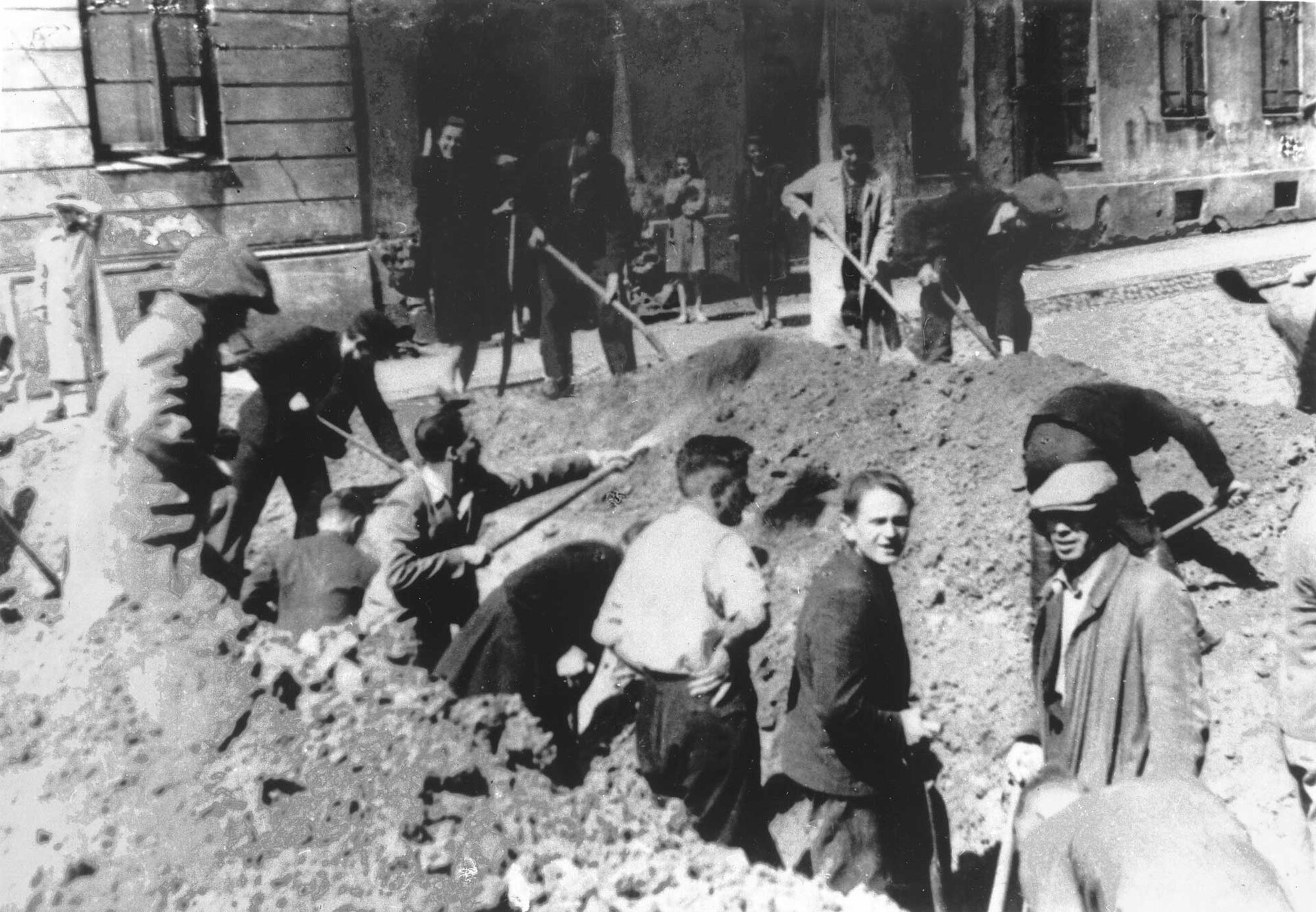 Zdjęcie przedstawia scenę kopania rowu przeciwczołgowego przez grupę dziesięciu mężczyzn i dwóch kobiet, którym przyglądają się ludzie stojący obok wykopu oraz w bramie budynku widocznego na drugim planie. Kopiący nie są ubrani w strój roboczy, a codzienny: mężczyźni mają koszule i marynarki, a kobiety noszą na sobie sukienki i płaszcze. 