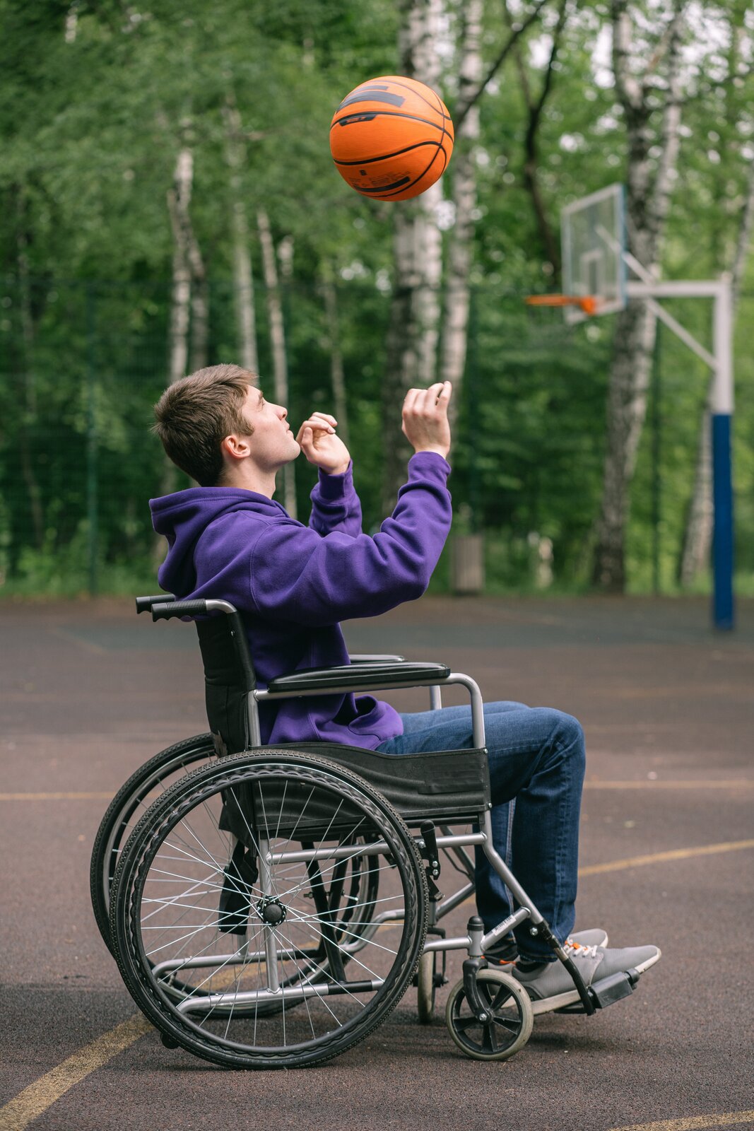 Zdjęcie przedstawia młodego mężczyznę na wózku. Mężczyzna znajduje się na boisku do koszykówki. Podrzuca do góry piłkę go koszykówki.