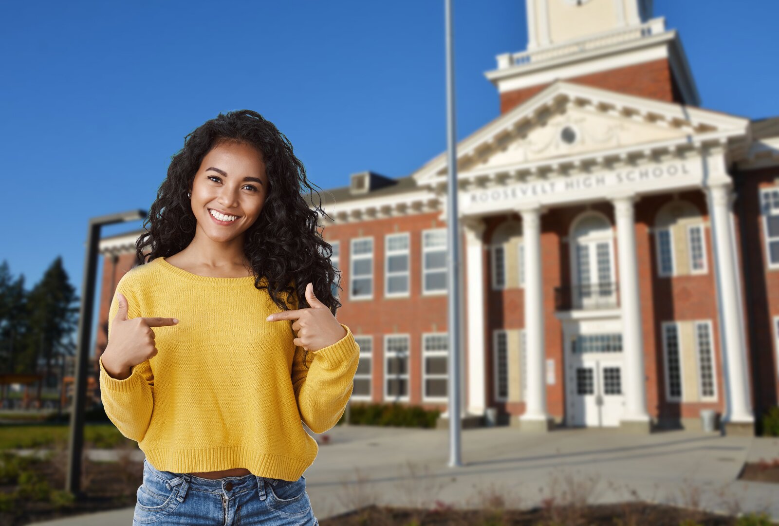 Zdjęcie przedstawia młodą dziewczynę, uchwyconą od bioder w górę, stojącą przed budynkiem widocznym w tle. Dziewczyna jest uśmiechnięta, ma długie, ciemne, rozpuszczone włosy i ubrana jest w żółty sweter oraz dżinsy. Jej ręce są zgięte w łokciach i uniesione, a palce wskazujące wyprostowane, układając się w gest wskazania na siebie. W tle znajduje się monumentalne wejście do budynku, wyróżniające się czterema wysokimi kolumnami zwieńczonymi tympanonem – trójkątnym szczytem. Po lewej stronie widoczne są dwa rzędy dużych okien.