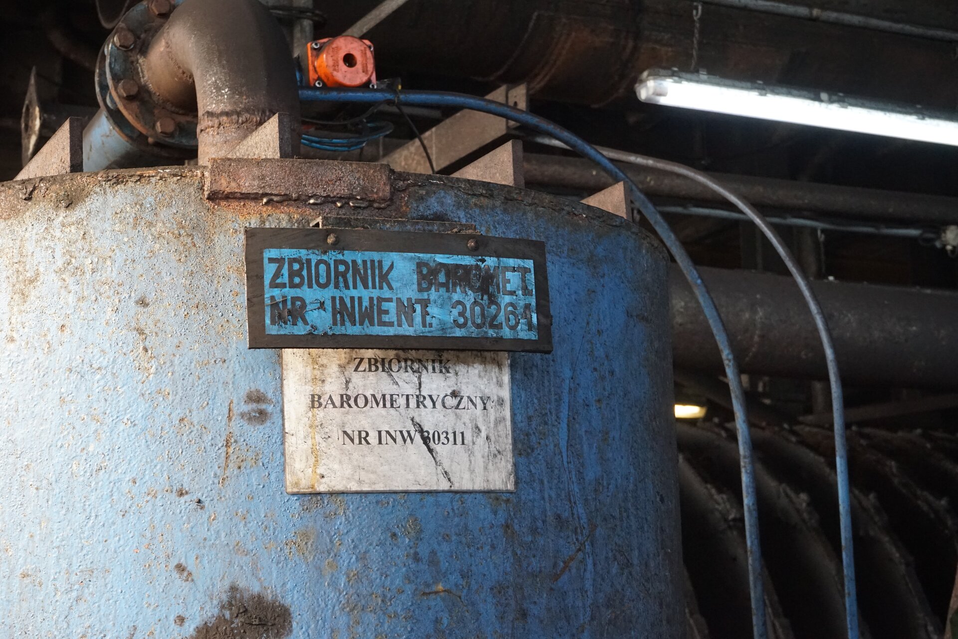 Zdjęcie przedstawia zbiornik barometryczny. Ma on cylindryczny kształt i jest zorientowany pionowo. Na nim znajduje się tablica informacyjna z nazwą. W górnej części są poprowadzone przewody, transportujące sprężone powietrze. Zbiornik barometryczny jest elementem układu wykorzystywanym pomocniczo podczas pracy filtra próżniowego lub prasy filtracyjnej. Poniżej zdjęcia znajduje się przycisk multimedialny z nagraniem audio. Treść nagrania jest zgodna z tytułem zdjęcia.