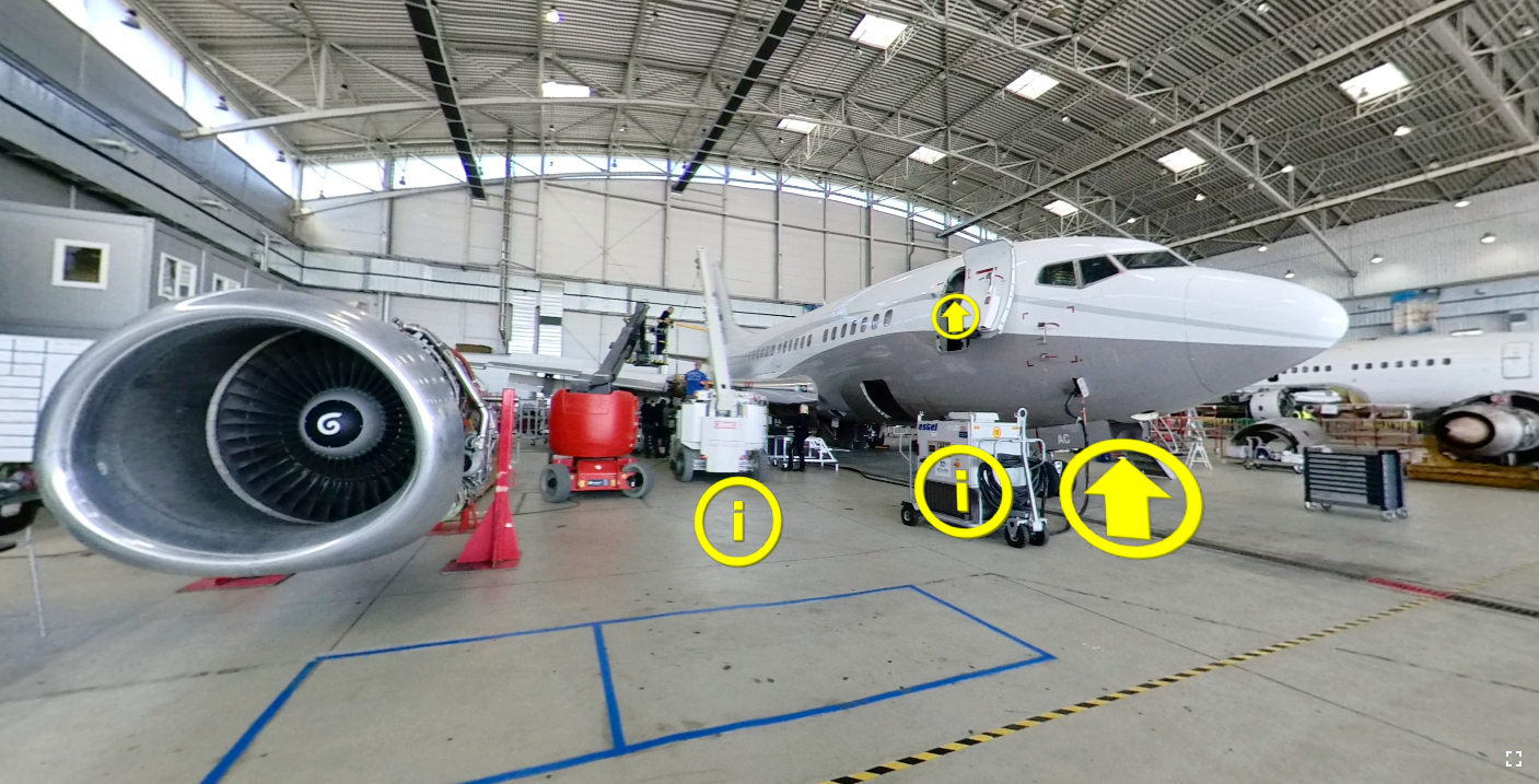 Ilustracja przedstawia przykładowy ekran wycieczki wirtualnej. Znajduje się na niej zdjęcie przedstawiające samolot podczas obsługi w hangarze. Samolot znajduje się po prawej stronie grafiki natomiast po lewej możemy zobaczyć zdemontowany silnik stojący na specjalnych stojakach. Dodatkowo na grafice znajduje się wiele maszyn i urządzeń wykorzystywanych podczas obsługi technicznej statków powietrznych. Na zdjęciu widoczne są również litery w okręgach, które oznaczają, że kryją się pod nimi informacje oraz strzałki w okręgu powodująca zmianę widoku. Oznaczenia ze strzałkami są przedstawione na drzwiach do samolotu jak i pod nim, z kolei kółka z literami znajdują się na urządzeniu stojącym przy samolocie oraz na podłodze hangaru.