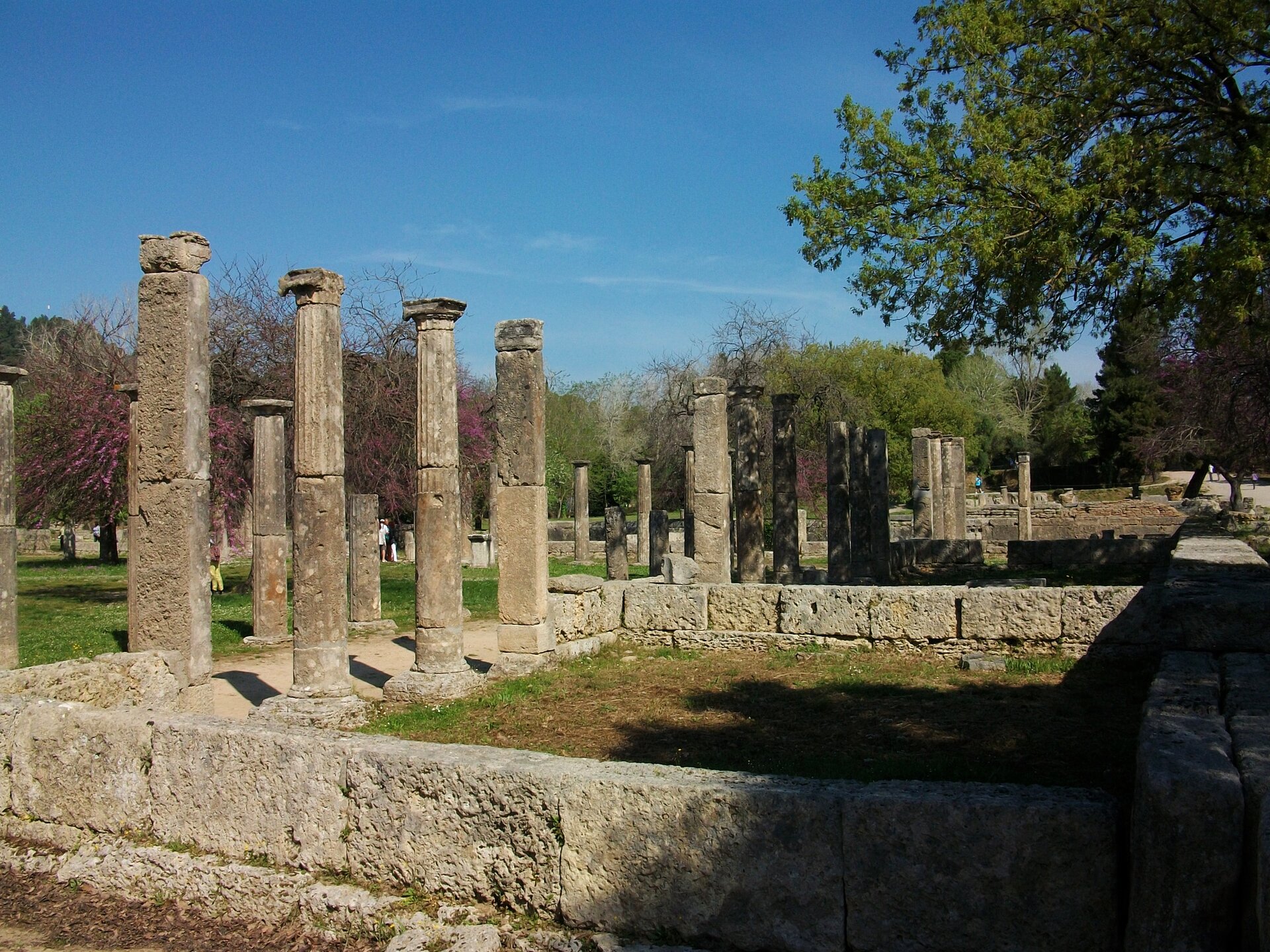 Zdjęcie przedstawia widok na dwa szeregi kolumn, ustawionych od lewej krawędzi fotografii ku centrum, pod nimi dolna część konstrukcji ściany budynku i nieregularnie rozrzucone głazy. Fragmenty budynku w centralnej części fotografii. Zdjęcie wykonane za dnia, przy wykorzystaniu naturalnego światła słonecznego, padającego z lewej strony ku centrum fotografii. Po prawej stronie widoczny zagajnik z pochylonymi ku centrum fotografii drzewami. 