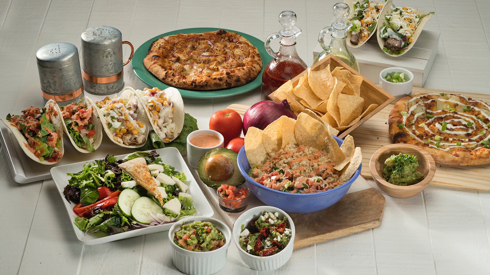 Zdjęcie przedstawia blat , na którym ustawiono przygotowane dania. Widoczne są dwie pizze na osobnych talerzach, taco na półmisku, sałata z ogórkiem papryką i oliwkami, chipsy z sosami w miseczkach. Obok postawiono w karafkach oliwę i ocet balsamiczny oraz solniczkę i pieprzniczkę.