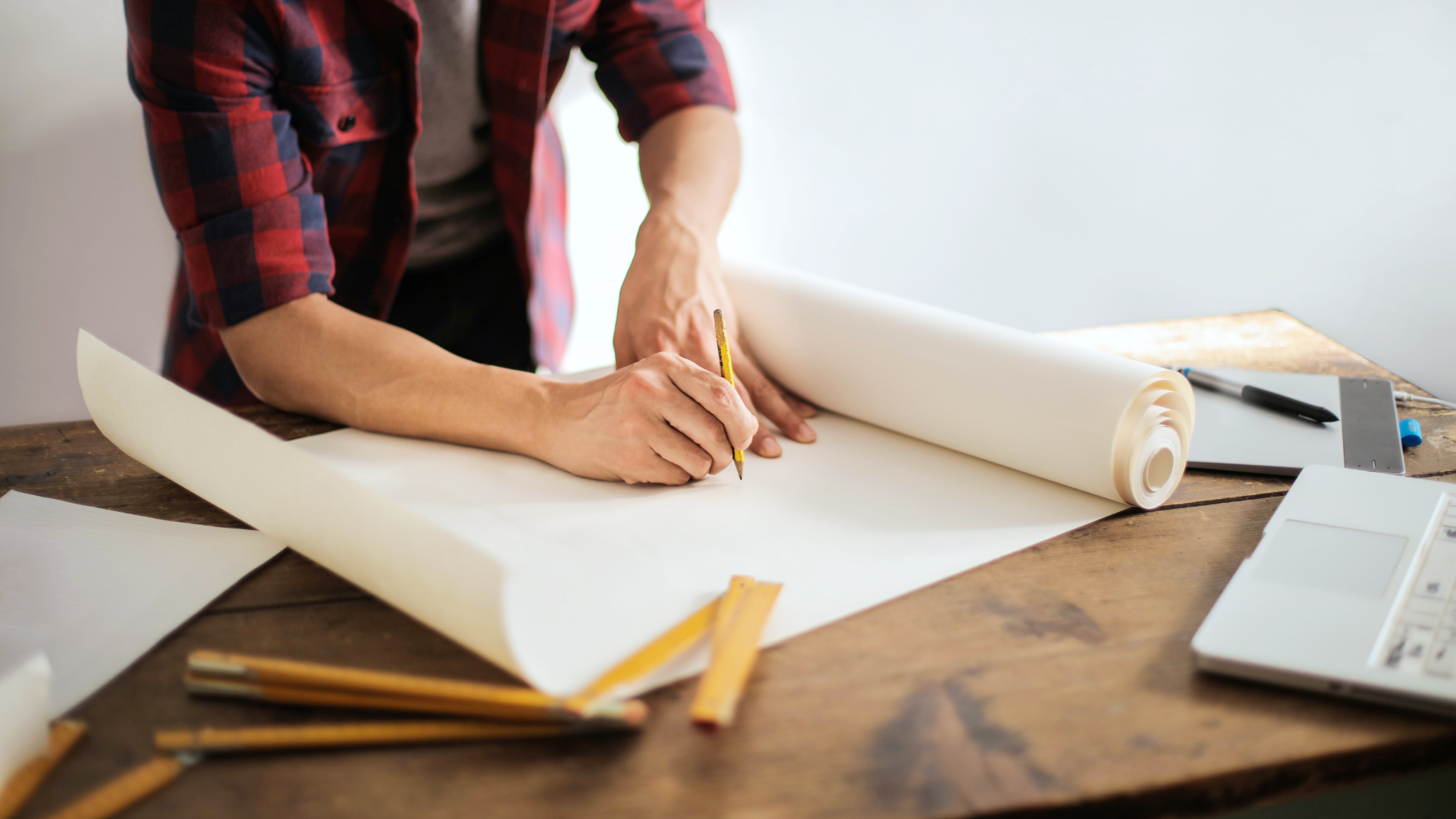 Zdjęcie przedstawia ręce mężczyzny, który trzyma w ręku ołówek i zamierza coś rysować na rozłożonym na stole kartonie.