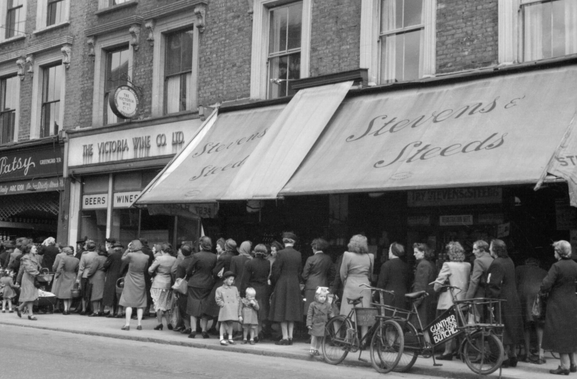 Zdjęcie przedstawia londyńską ulicę w 1945 roku. Przed sklepem, na chodniku stoi długa kolejka kobiet. Nieopodal nich widać kilkoro dzieci.