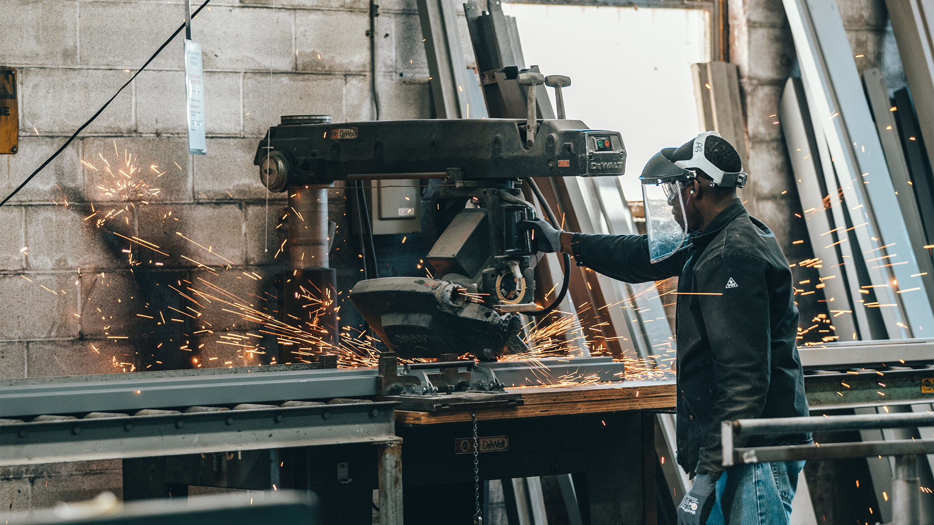 Na grafice przedstawiono proces spawania. Przy spawarce sporych rozmiarów stoi obsługujący ją spawacz. Z maszyny lecą iskry, będące częścią procesu spawania.