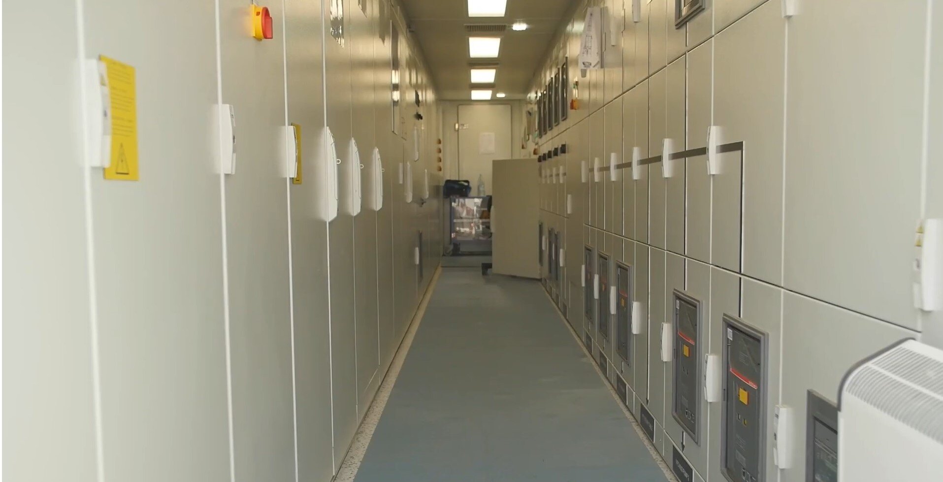 Zdjęcie przedstawia prądową rozdzielnie sterującą. Jest to pomieszczenie z białymi zamkniętymi szafami po obu stronach