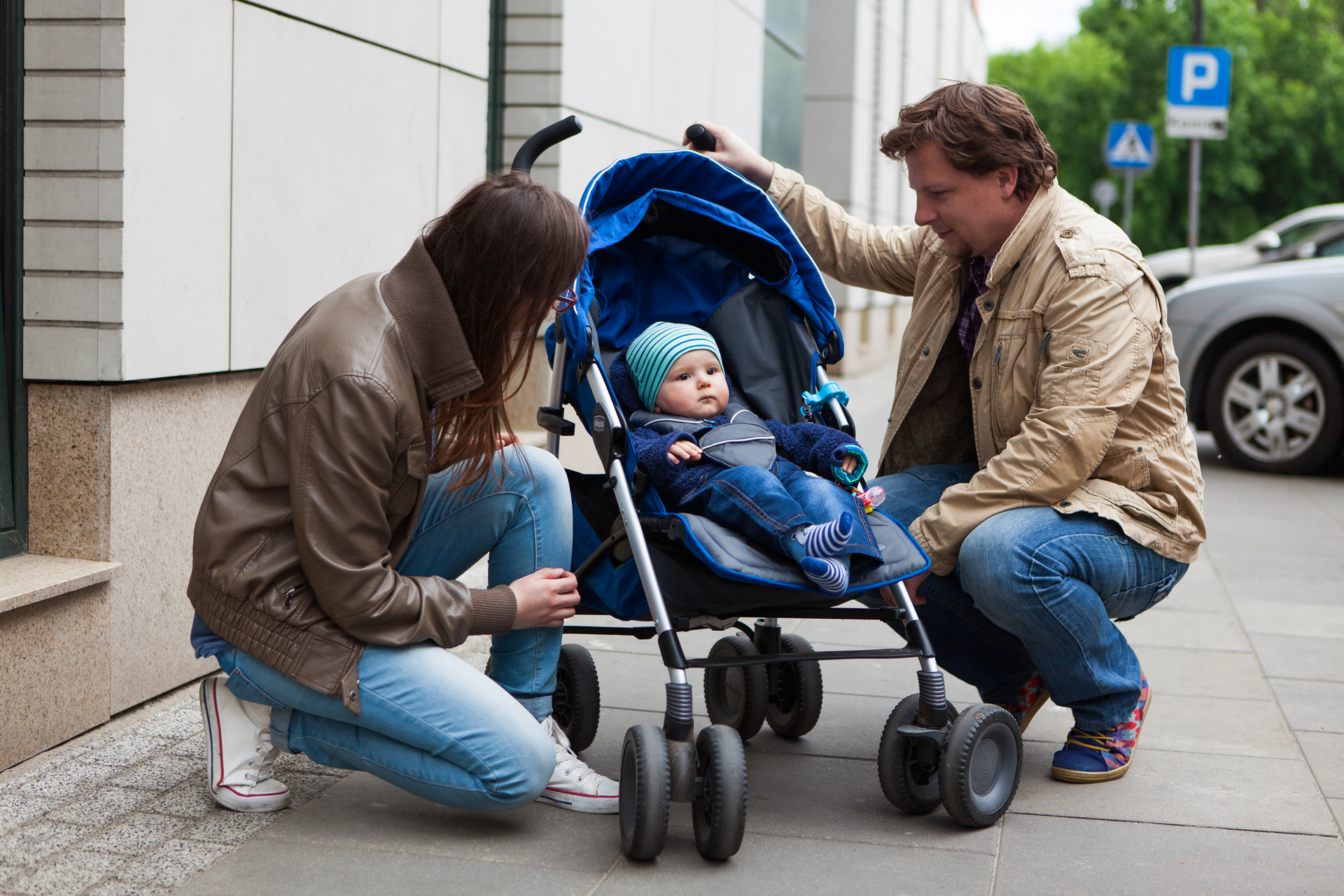 Fotografia przedstawia parę ludzi na ulicy w wieku około 30 lat, pochylonych nad wózkiem z małym dzieckiem. W tle znajdują się zaparkowane przy chodniku samochody oraz ściana budynku.