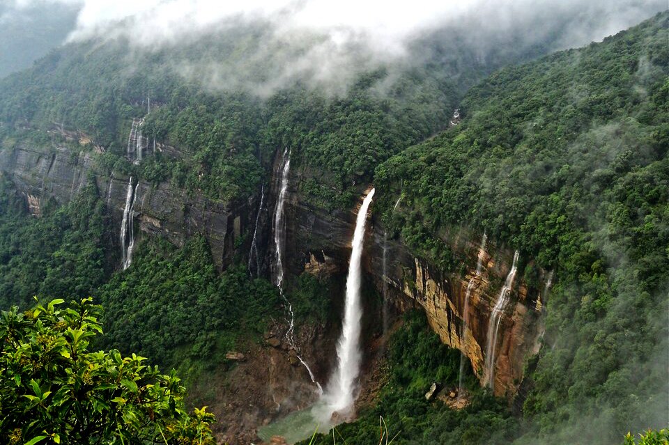 Fotografia barwna przedstawiająca Wodospad Nohkalikali w pobliżu Czerapuńdżi, miasta w północnych Indiach, najbardziej deszczowego miejsca na Ziemi. Fotografia przedstawia jeden duży i kilka mniejszych wodospadów. Wokół wodospadów, aż po horyzont rozciąga się las deszczowy. Zdjęcie wykonane zostało z lotu ptaka.