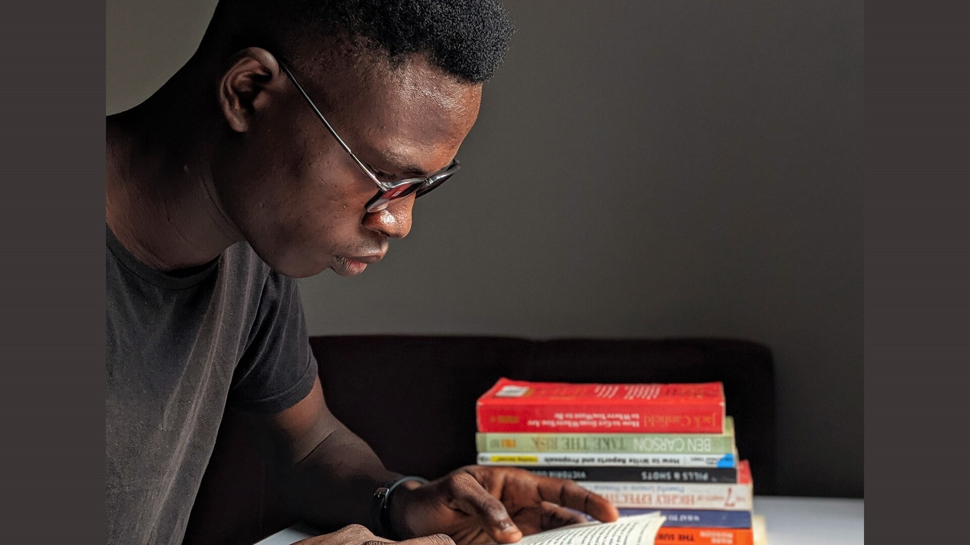 Zdjęcie przedstawia siedzącego przy biurku mężczyznę, od pasa w górę, w ujęciu z prawego boku. Mężczyzna ma krótkie czarne włosy, ubrany jest w brązową koszulkę z krótkim rękawem i ma okulary. Skupiony pochyla się nad książką, którą trzyma w dłoniach. Na lewo od niego widoczny jest stos książek leżących na biurku. 