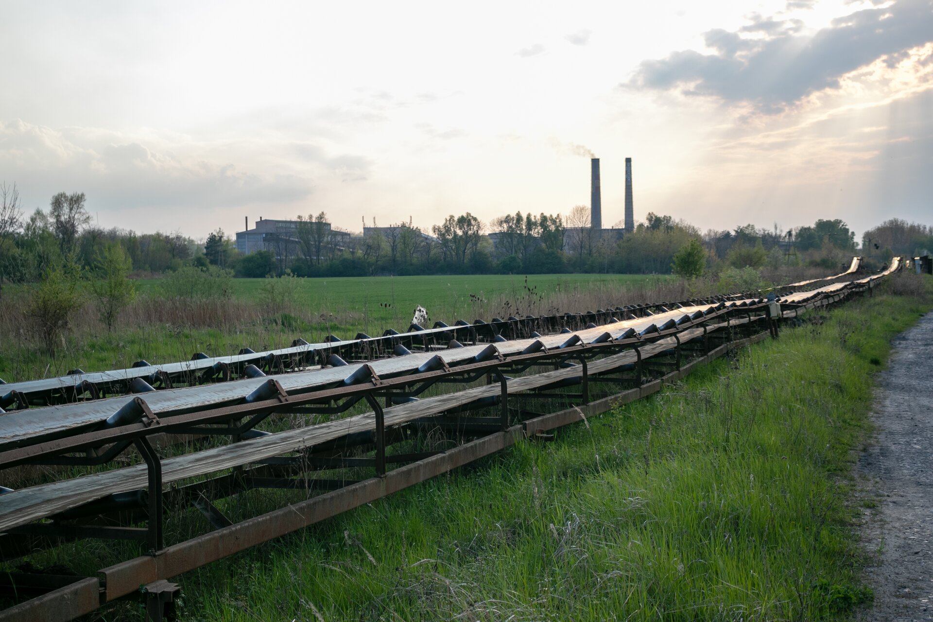 Na zdjęciu widoczny jest przenośnik taśmowy wykorzystywany do transportu materiału na większe odległości.Przenośnik jest układem modułowym. Na lekko nachylonych rolkach o kształcie walca osadzona jest taśma. Po lewej stronie przenośnika widoczny jest identyczny układ. Oba urządzenia biegną przez polanę. Po lewej stronie zdjęcia widać w oddali dwa wysokie kominy. Przenośniki biegną w ich stronę.Poniżej zdjęcia widoczny jest przycisk multimedialny z nagraniem audio. Treść nagrania jest zgodna z tytułem zdjęcia.