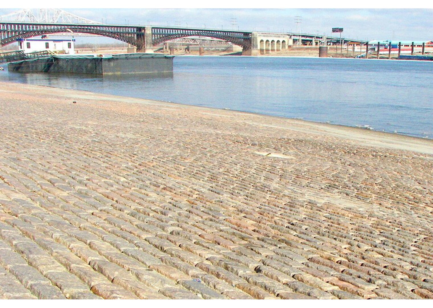  Zdjęcie przedstawia szeroki fragment brzegu wykonany z kostki granitowej tuż nad rzeką. W oddali most oraz zabudowania.