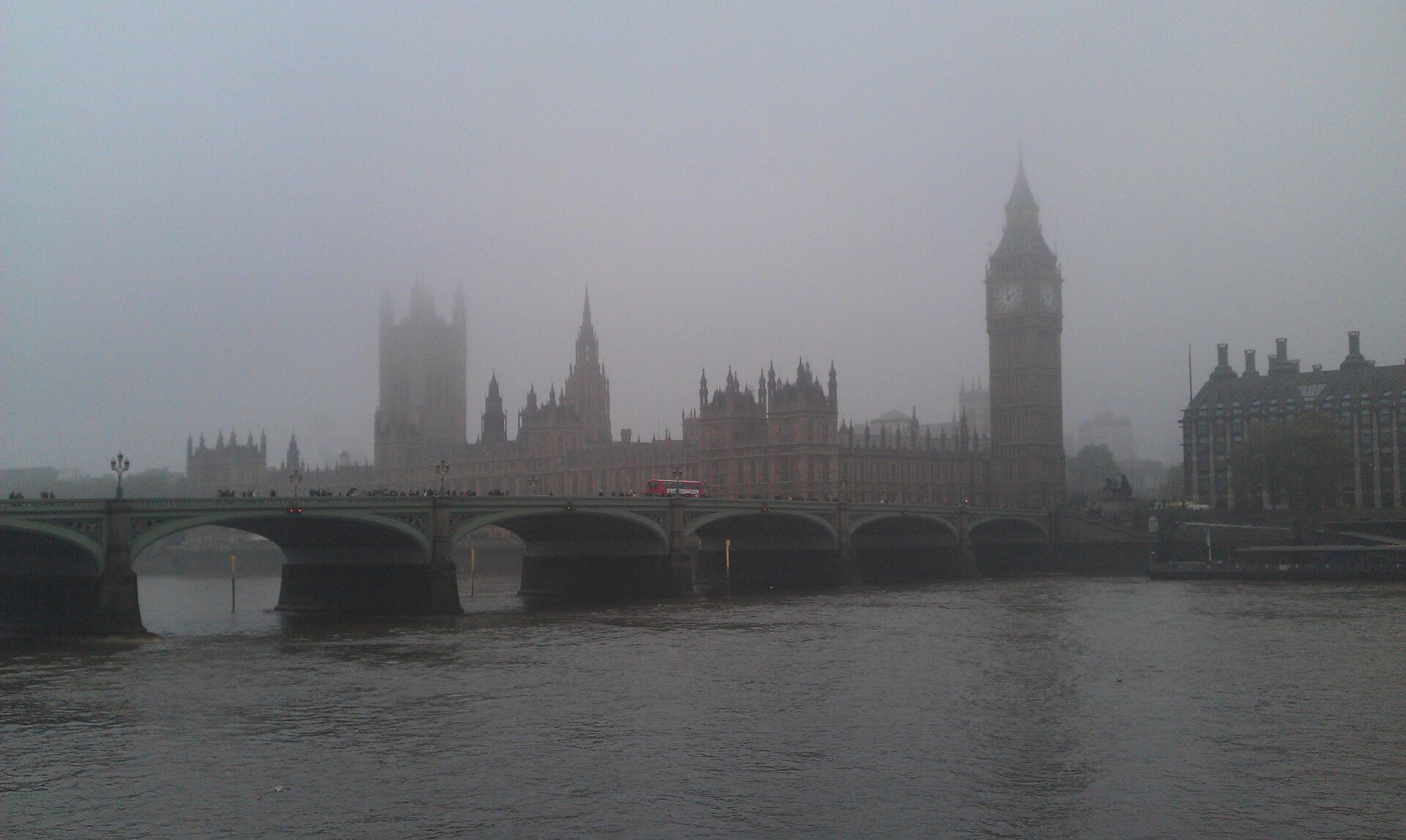 Zdjęcie przedstawia smog westminsterski w Londynie. Na pierwszym planie znajduje się Tamiza płynąca pod mostem. W tle są przysłonięte smogiem zabudowania miasta.