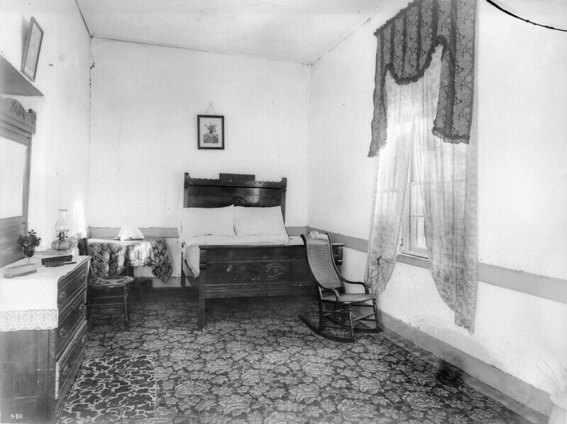 Zdjęcie przedstawia wnętrze sypialni. Stoi tu duże, drewniane, dwuosobowe łóżko, fotel bujany, bieliźniarka z lustrem oraz stolik i krzesło.