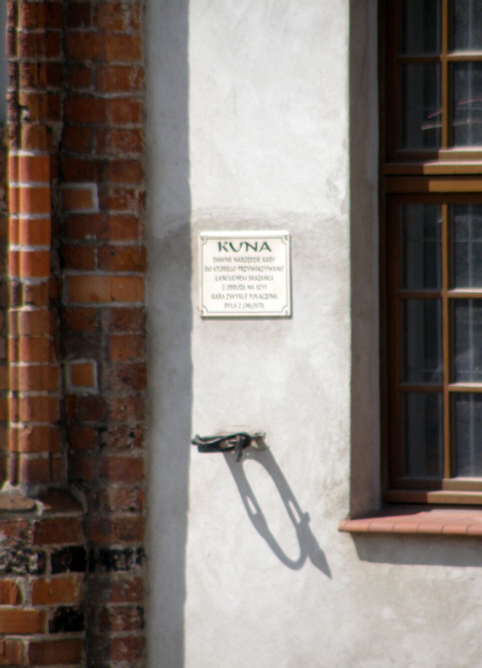 Zdjęcie przedstawia fragment ściany kamienicy. Do ściany kamienicy przymocowana jest żelazna obręcz. Nad obręczą jest tabliczka z informacją czym jest obręcz i do czego służyła w przeszłości.